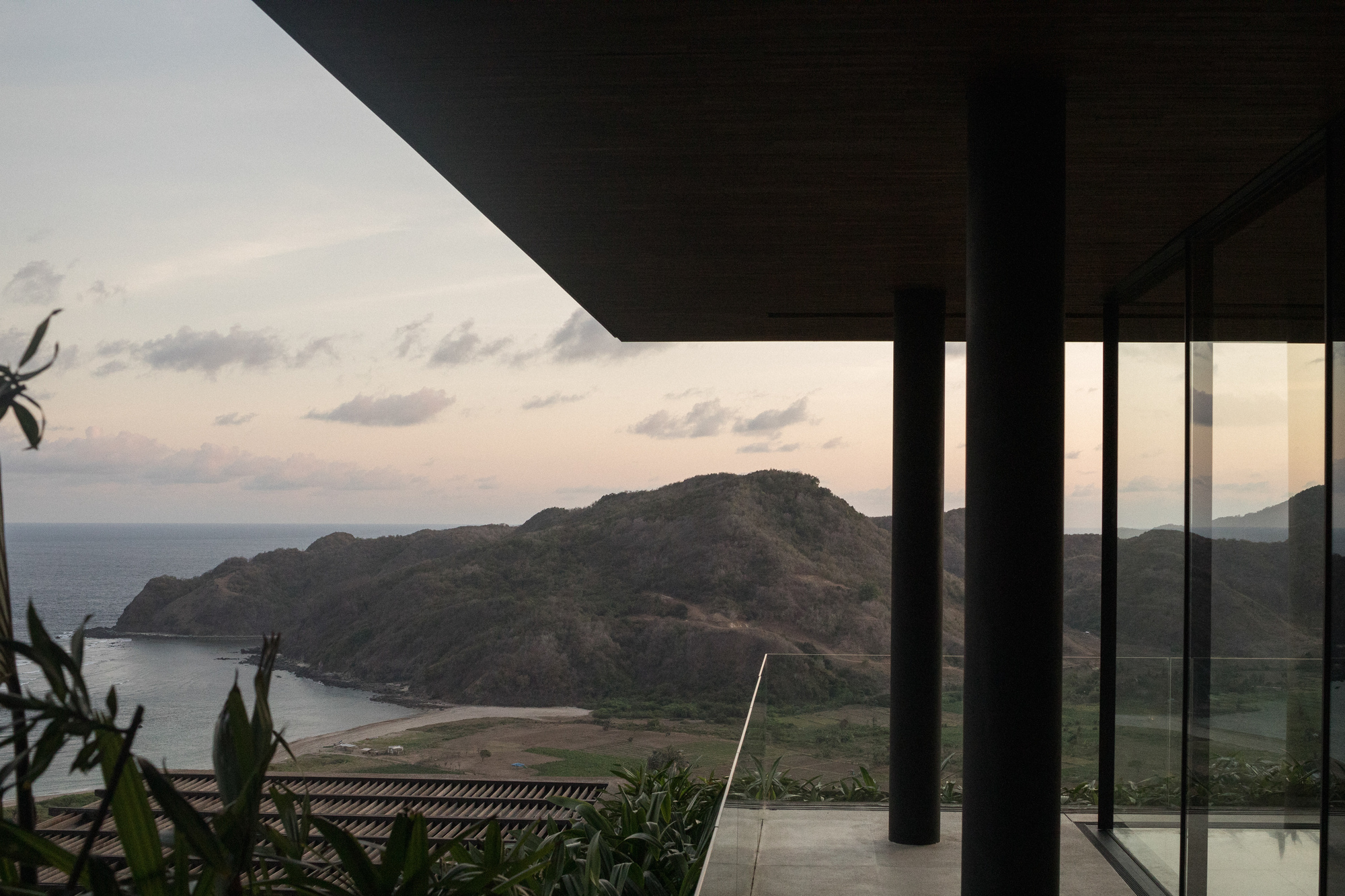 Dark roof overhang and round steel column framing a green headland and bay at dusk with pink-tinged clouds