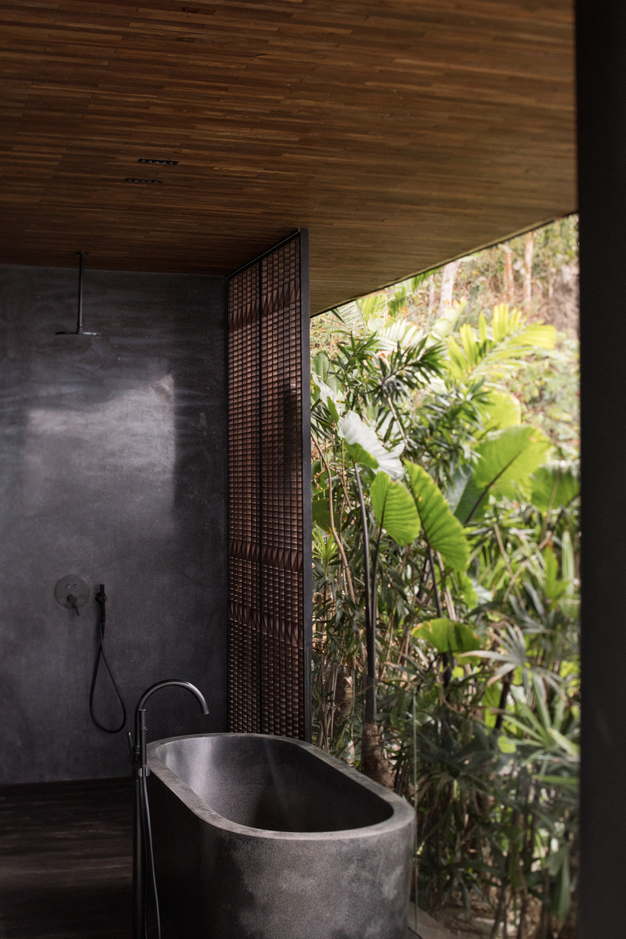 Stone bathtub beside a concrete-finish wall and rain shower, with a lattice screen and jungle foliage under a timber ceiling