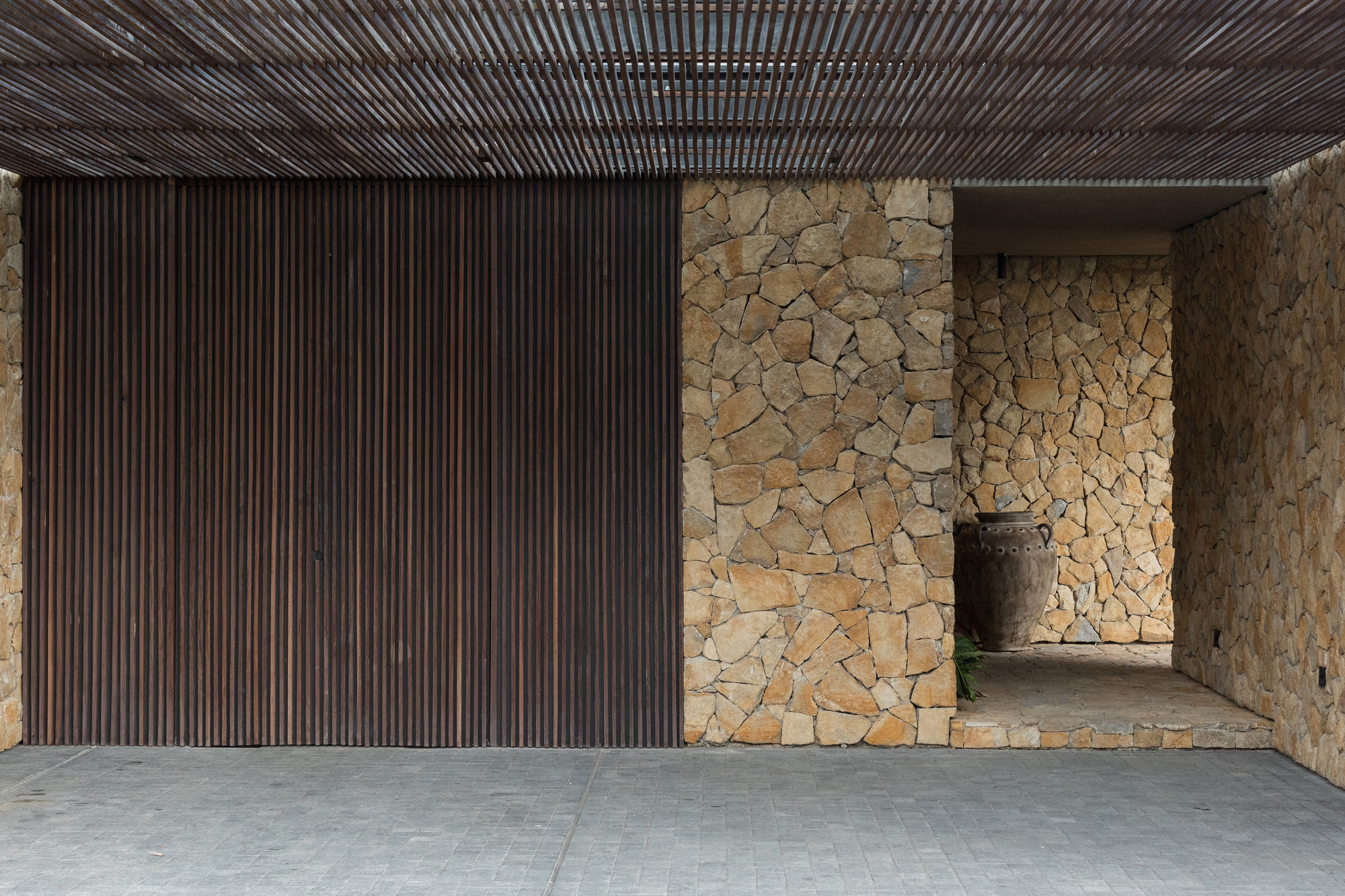 Carport entrance with dark vertical timber slats, rubble stone walls and a clay urn beneath a slatted timber ceiling
