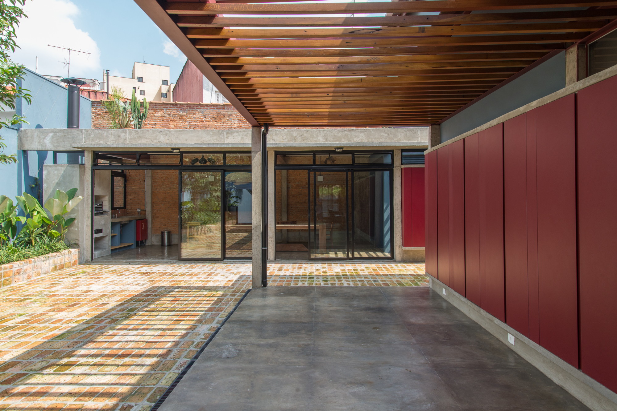Covered terrace with slatted timber roof, red panel wall and glazed doors opening onto a brick-paved court in sunlight
