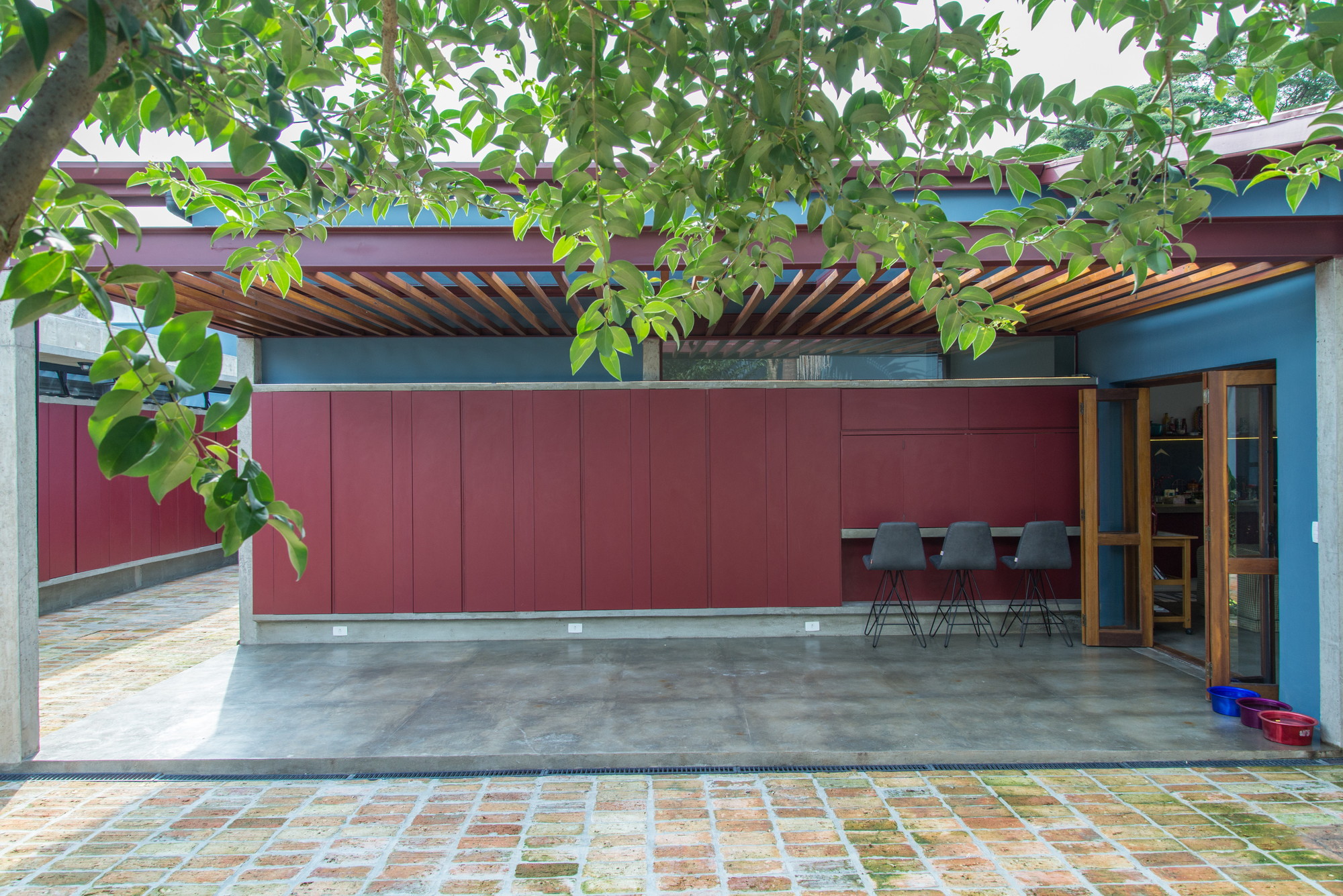 Red panel wall and outdoor bar stools under a timber slat roof, framed by ficus leaves in daylight