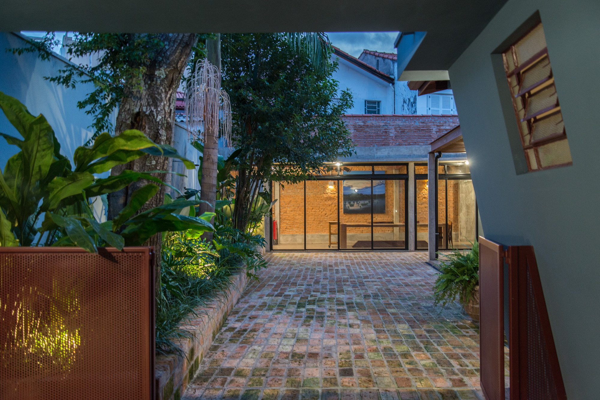 Reclaimed brick paved courtyard leading to a glazed pavilion with exposed brick walls, lit at dusk among tropical plants