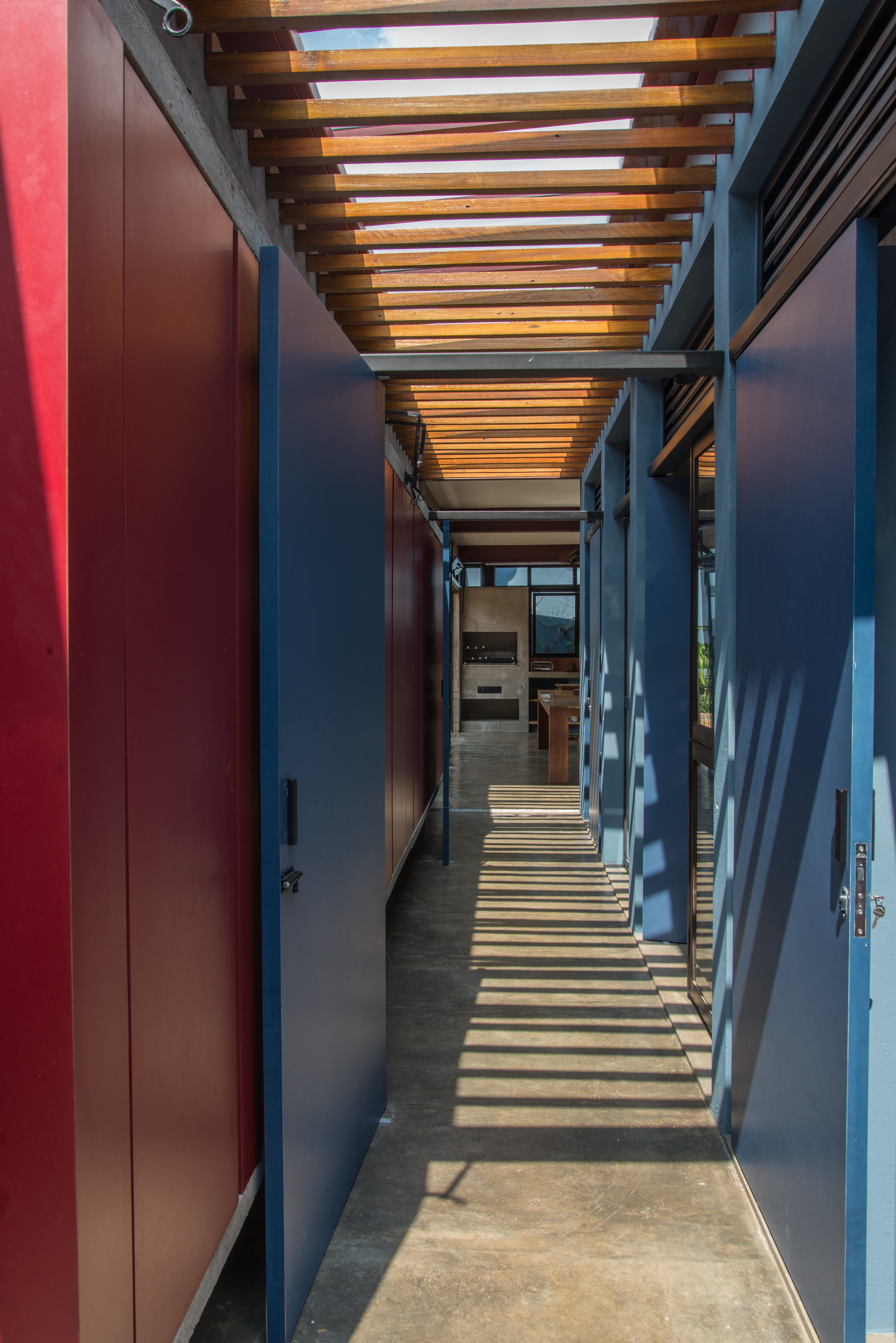 Narrow concrete corridor between red and blue panel doors, with a timber slat canopy casting striped shadows across the floor