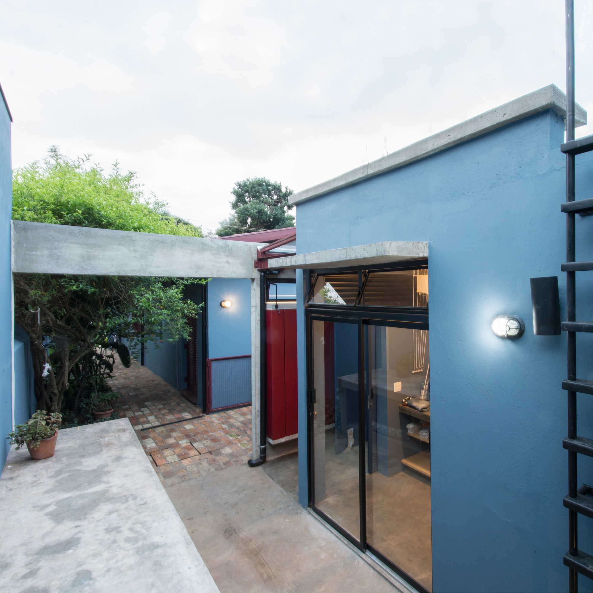 Blue rendered volume with sliding glass doors and a concrete lintel beside a brick path under an overcast sky