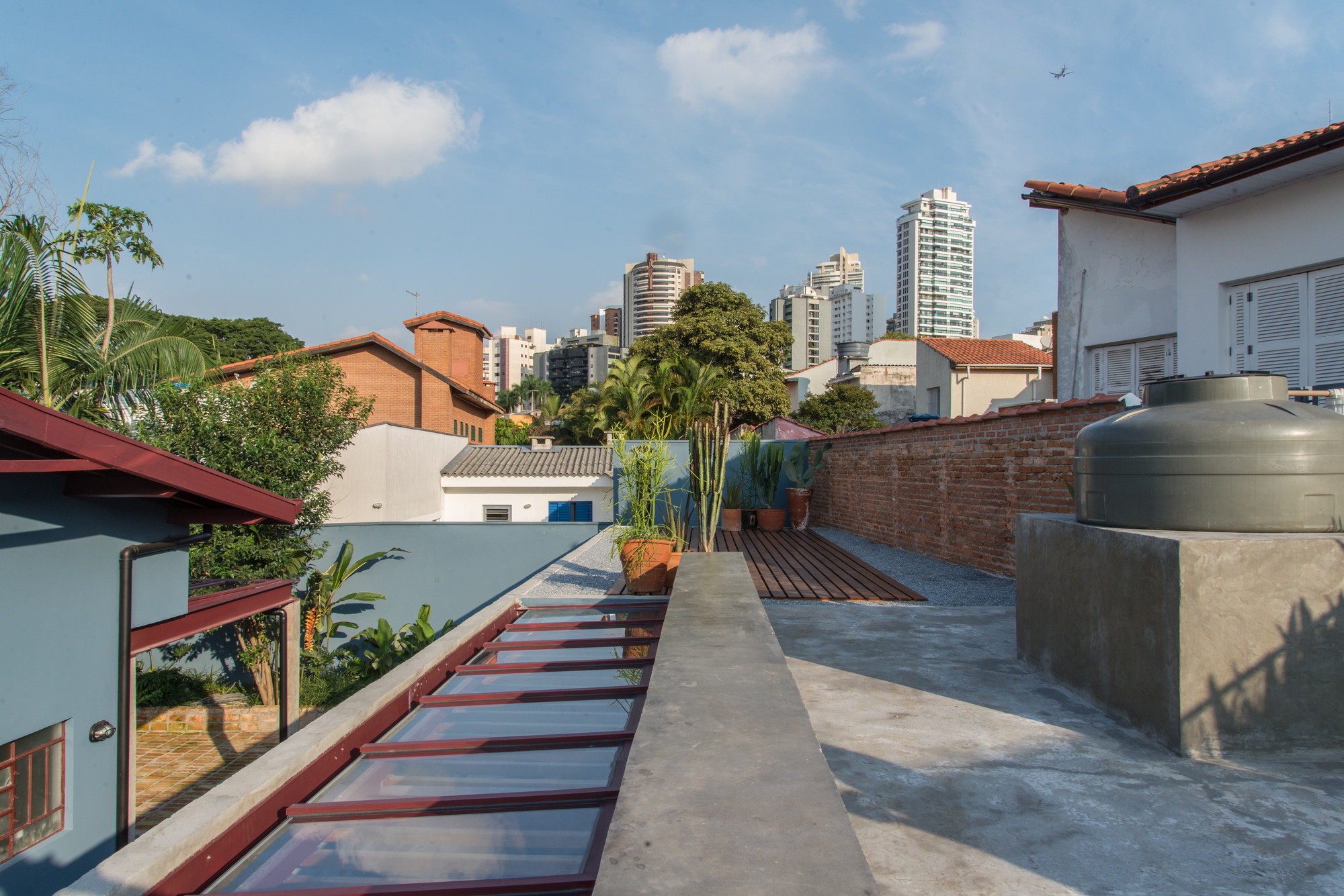 Rooftop terrace with a red-framed skylight, concrete water tank and timber deck, set against distant apartment towers in blue sky
