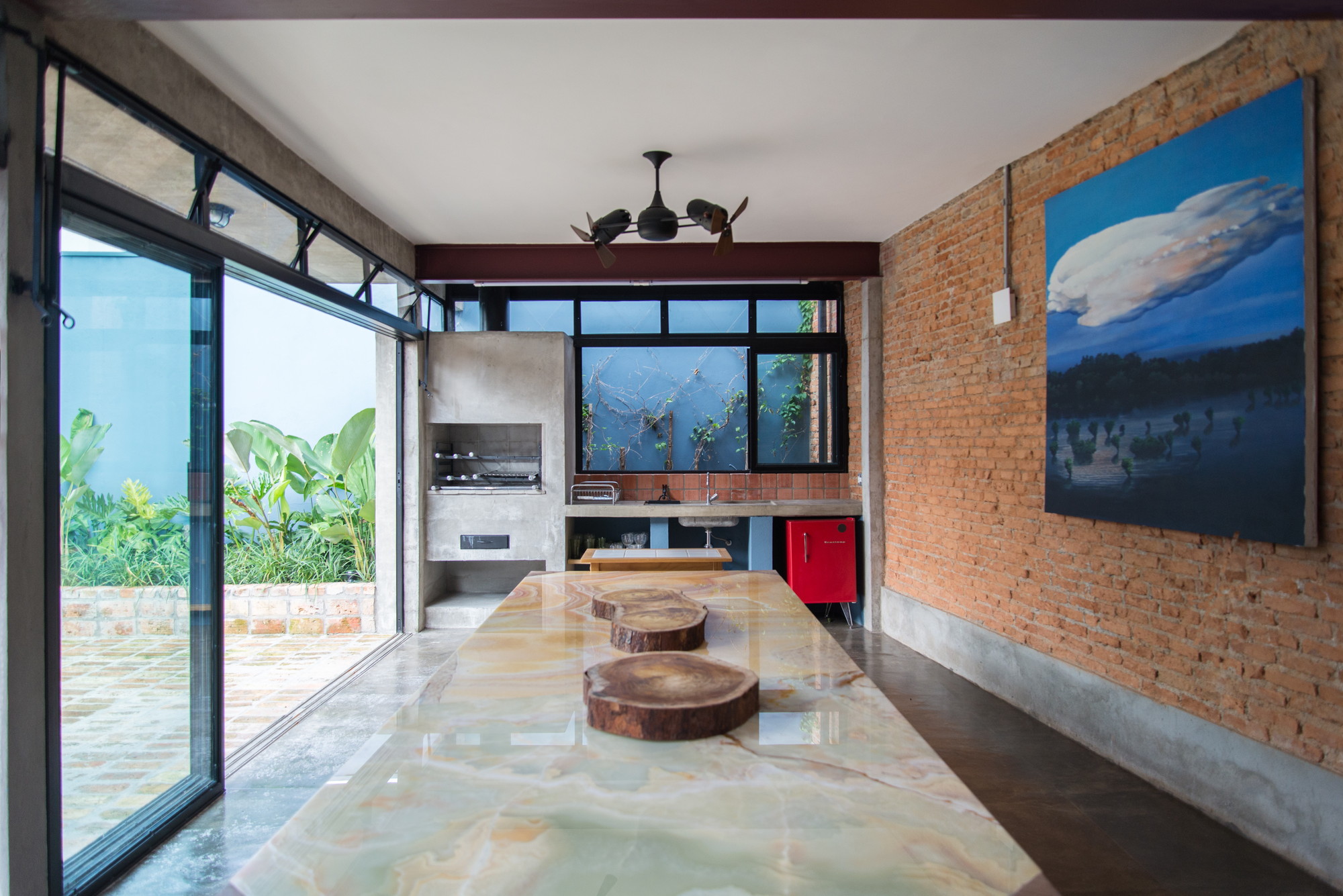 Open dining pavilion with an onyx-topped table, concrete barbecue and exposed brick wall hung with a large cloud painting