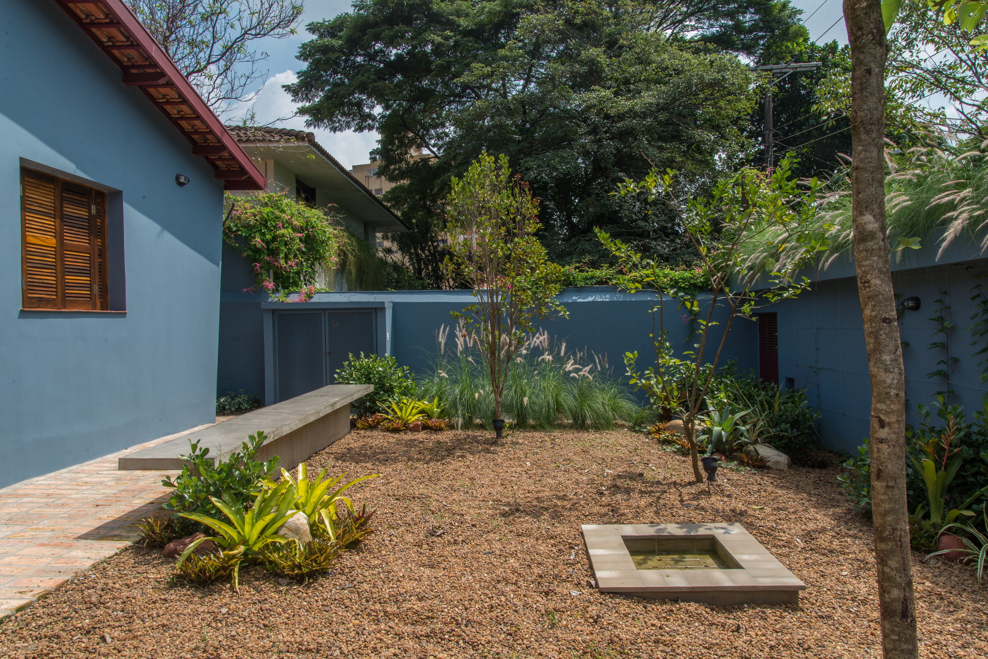 Gravel courtyard with a small square water basin, concrete bench and bromeliads beside blue walls under tall trees in daylight