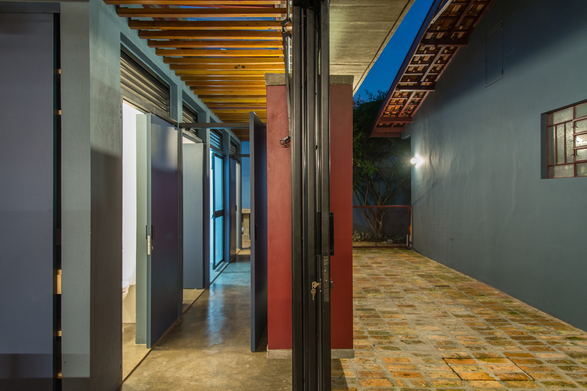 Covered passage with timber slat ceiling, red masonry pier and pivot doors opening onto a brick-paved court at dusk
