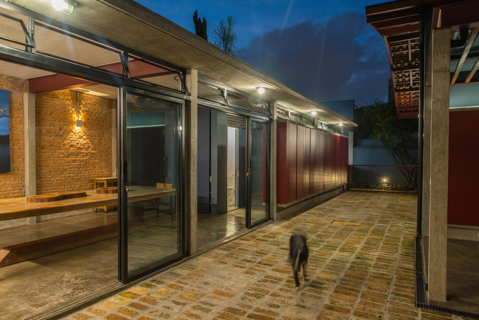 Glazed sliding doors open to a brick-walled room with a timber table, while a dog crosses the brick paving at night