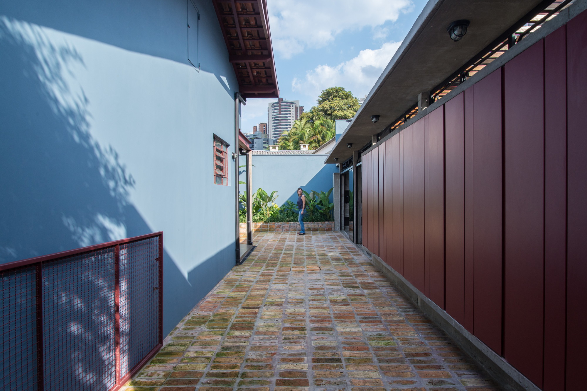 Narrow brick-paved passage between a blue wall and red panel cladding, with a person standing at the far end in sunlight