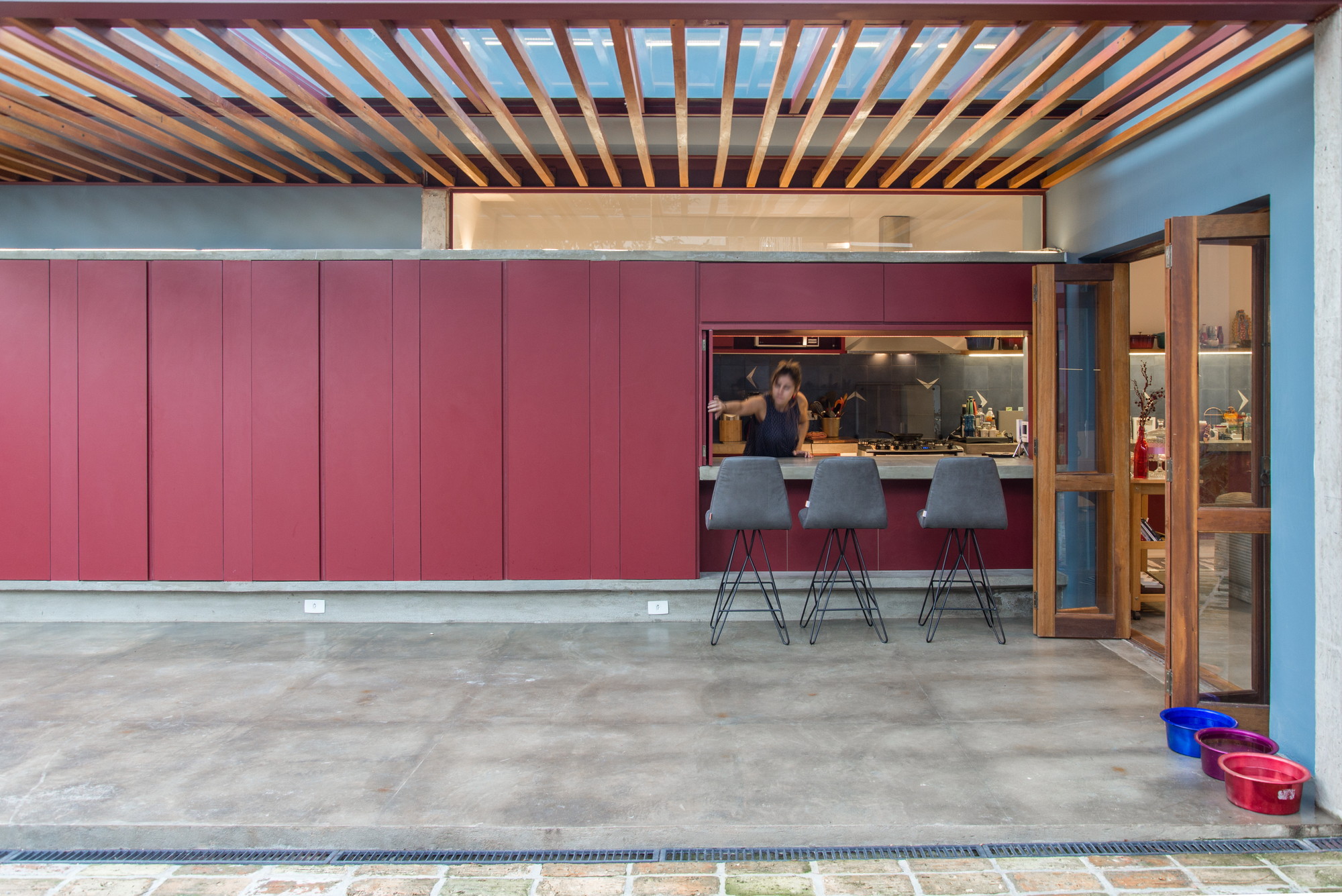 Kitchen pass-through in a red panel wall with three grey stools, a cook inside and a timber slat canopy above