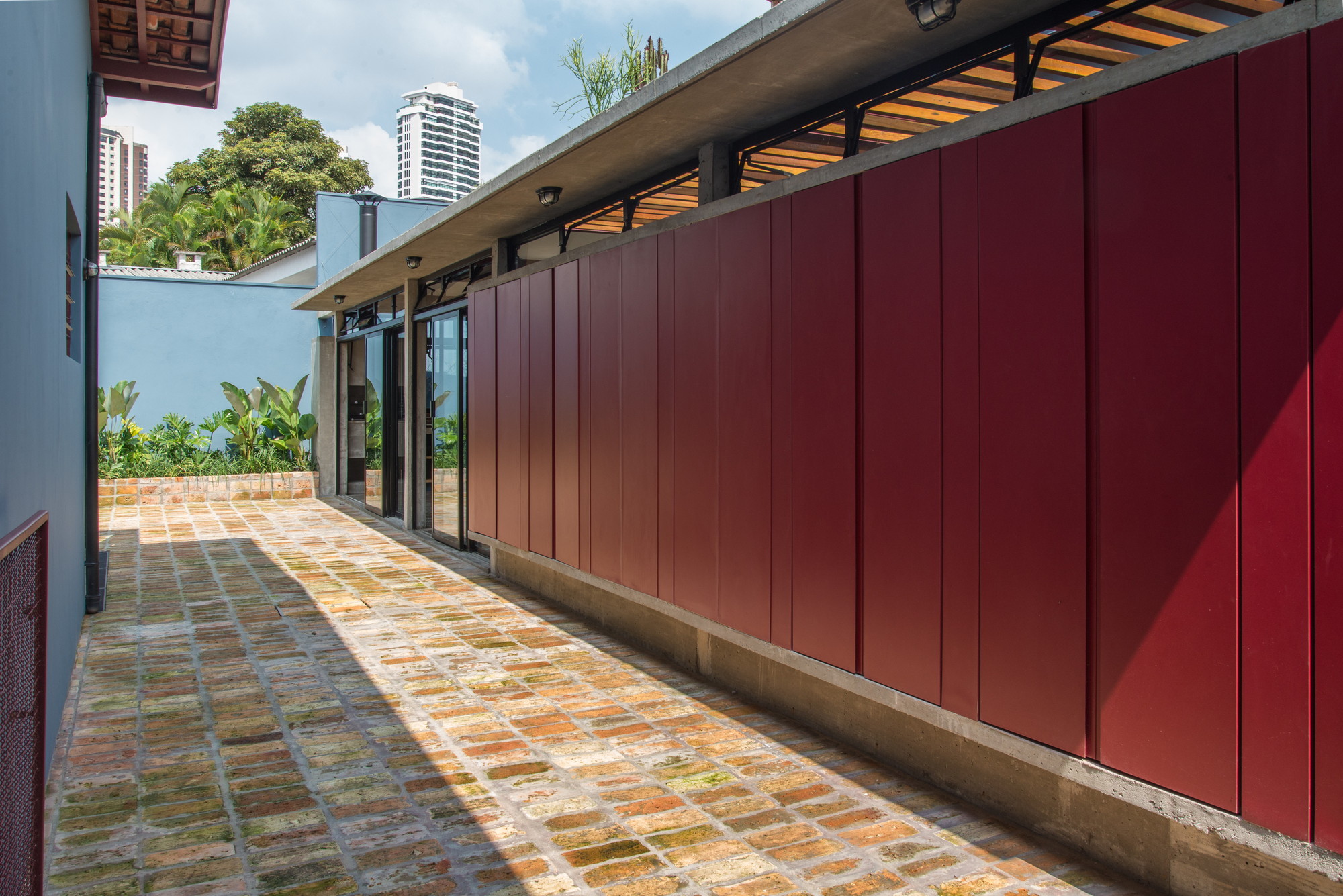 Side passage lined with a deep red panel wall and reclaimed brick paving, striped with afternoon sun and shadow