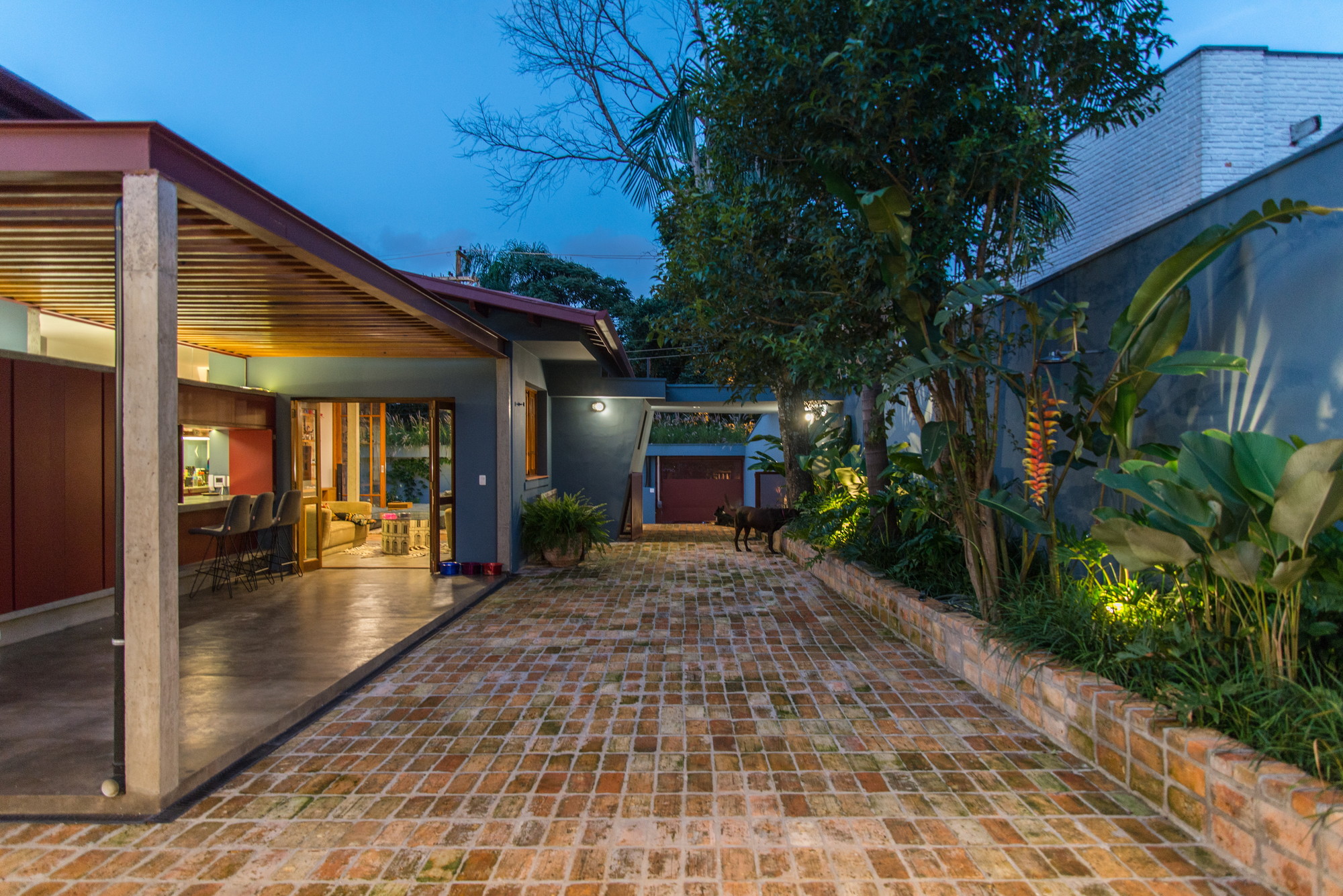 Brick-paved courtyard at dusk with a covered porch, glowing interior, planted bed and a dog near the far gate