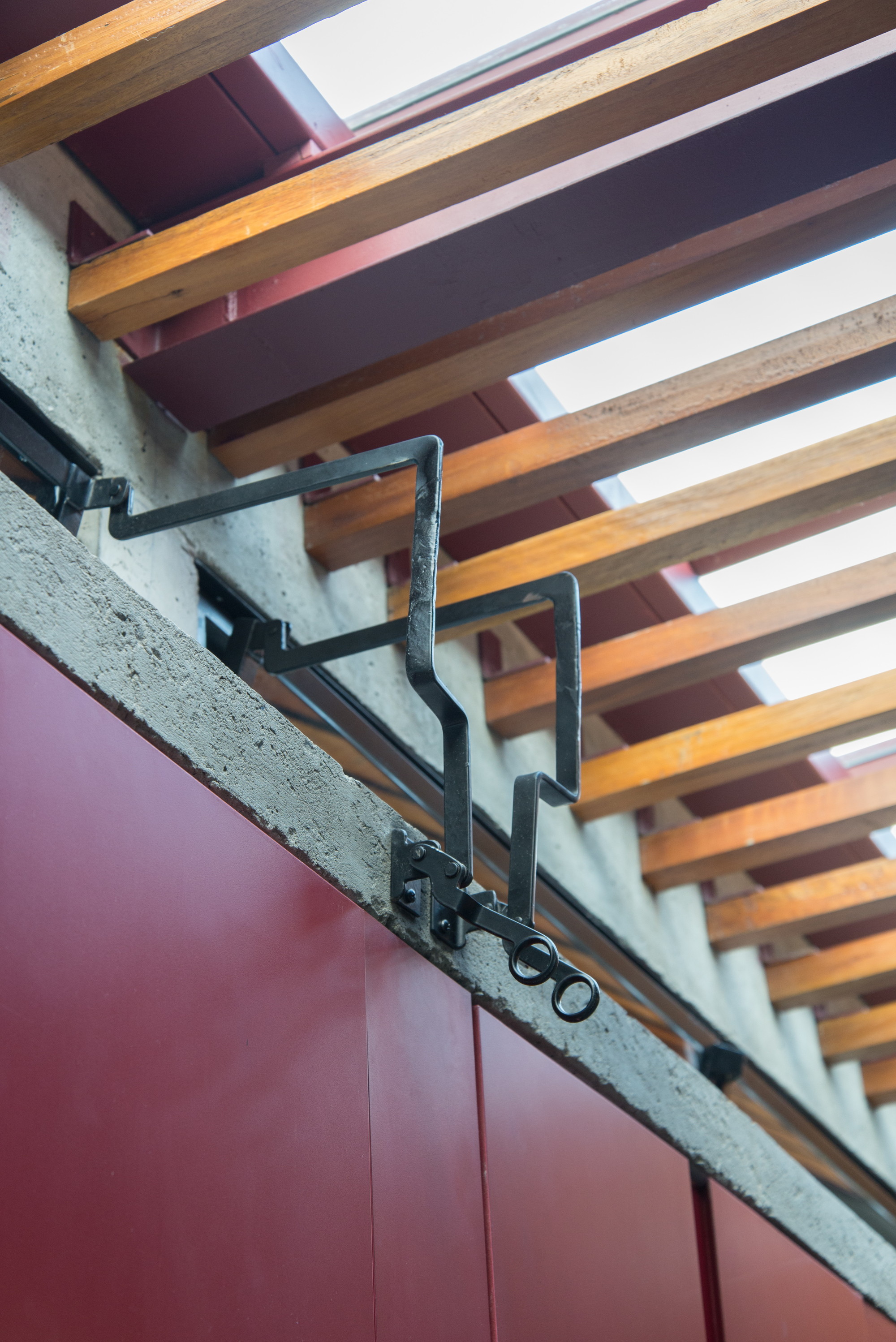 Close-up of a wrought iron window latch beneath timber rafters and red steel beams, with daylight coming through the roof