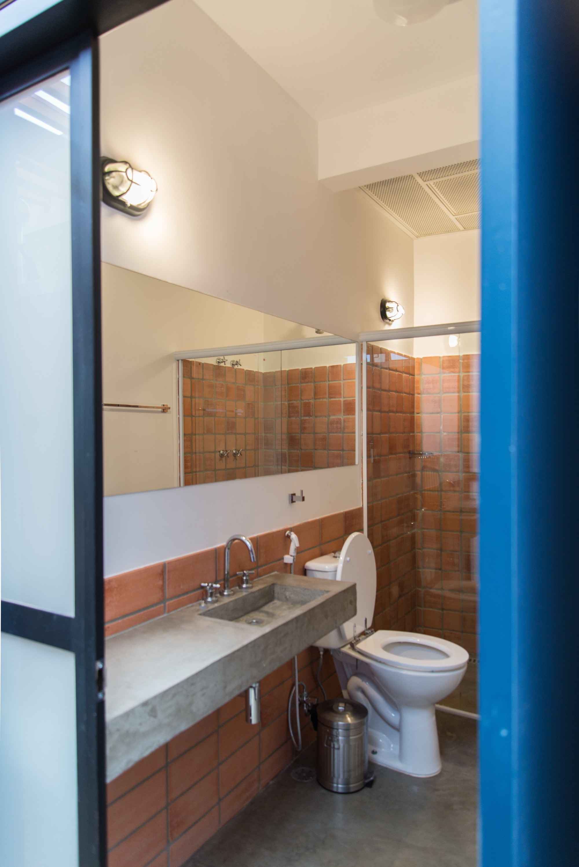 Bathroom seen through a blue-framed door, with a concrete trough sink, terracotta tile walls and a long mirror