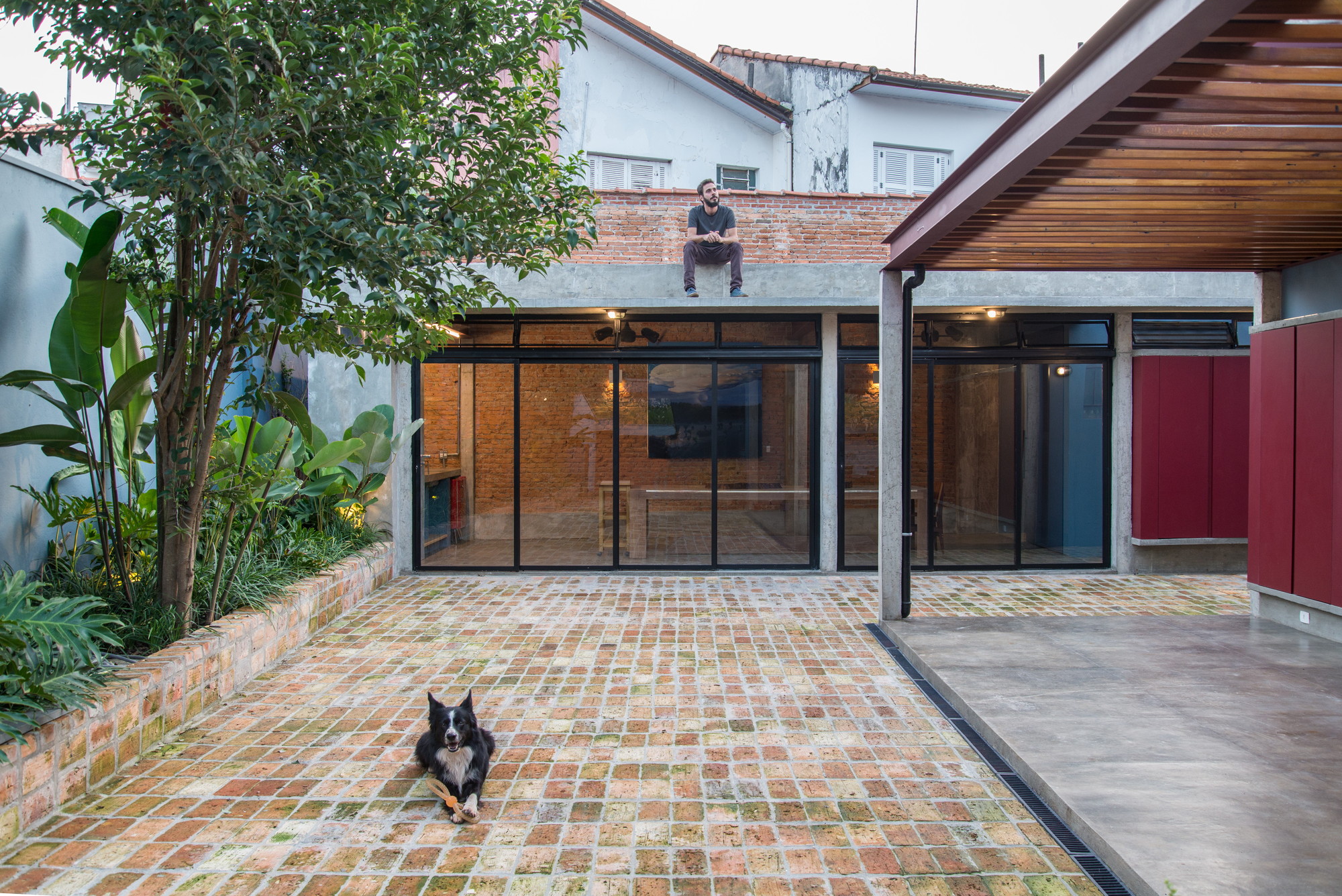 Brick-paved courtyard facing a glazed brick room, with a man seated on the roof edge and a dog lying in front
