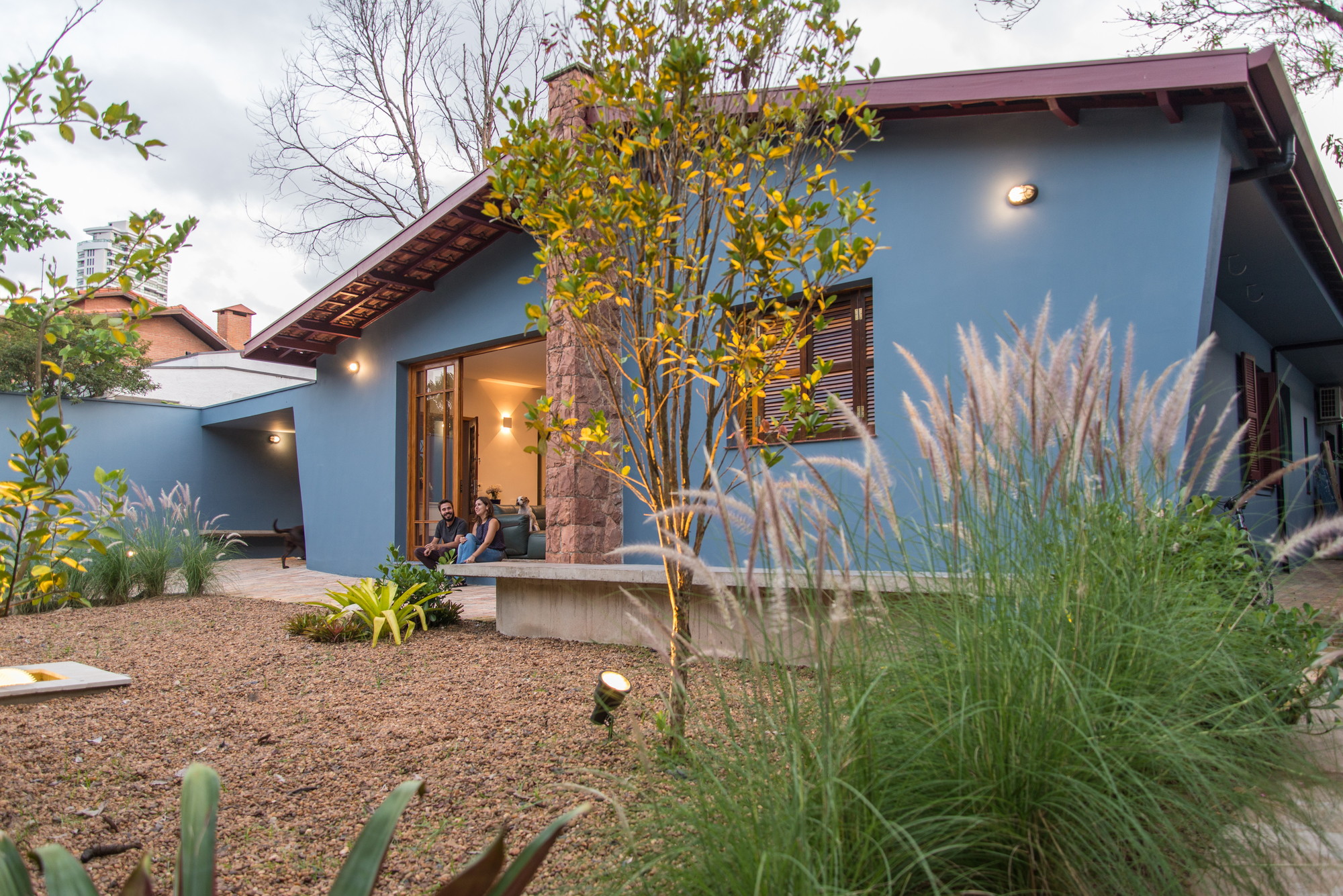 Blue stucco house with a sloped roof and stone column, seen through ornamental grasses and young trees at dusk