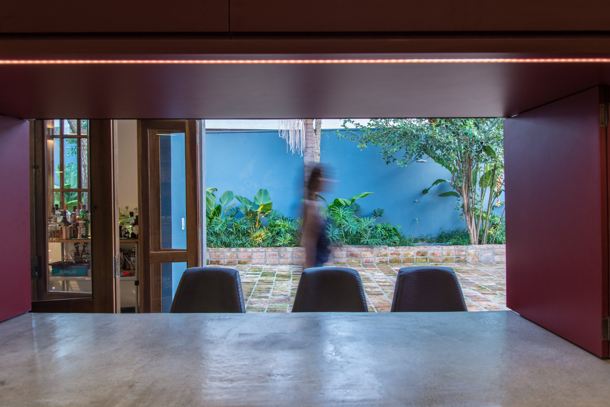 View through a red opening past a concrete counter and three chairs to a blue-walled garden, with a blurred figure walking by
