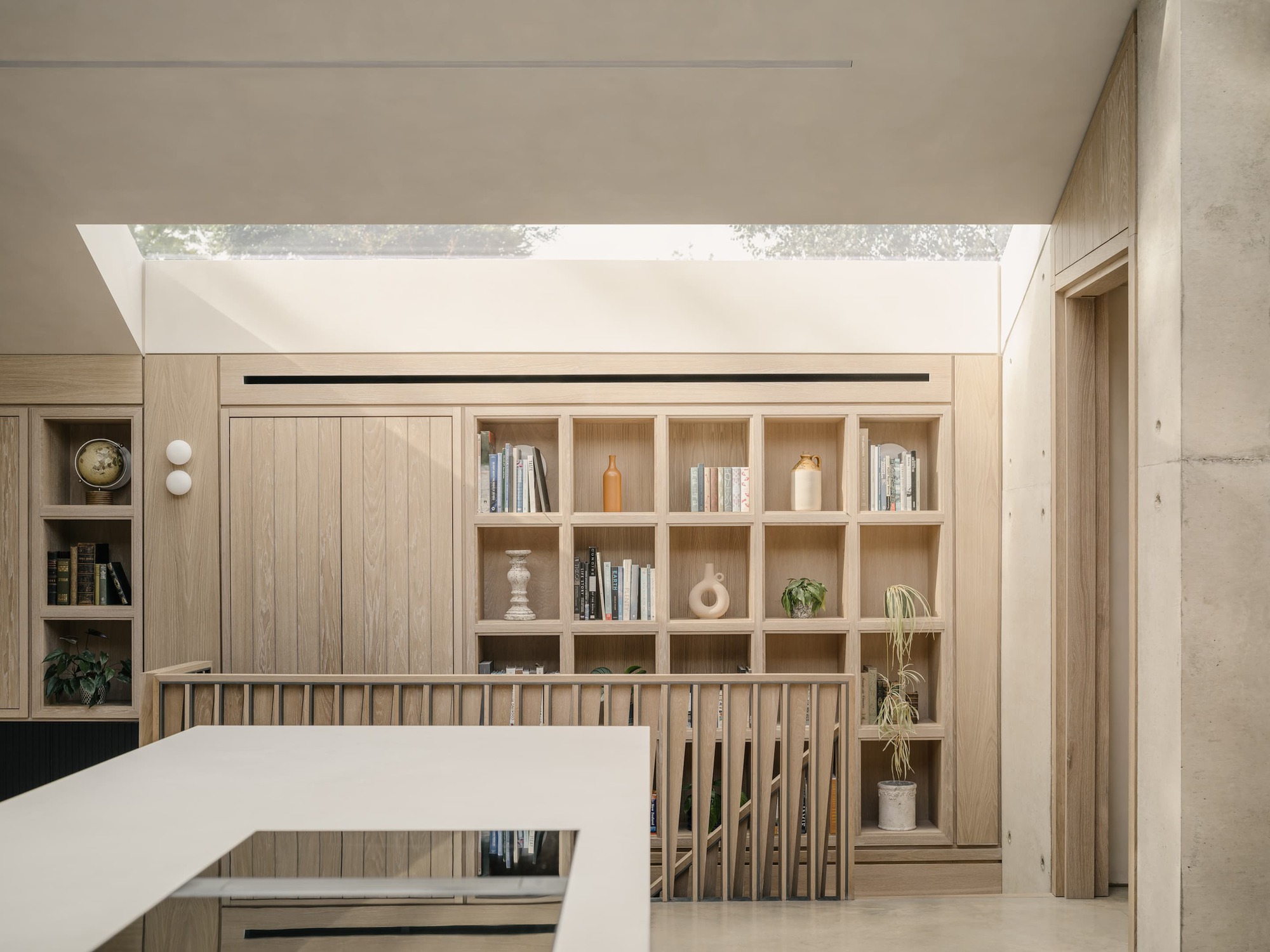 Oak bookshelf wall beneath a long rooflight, with a slatted balustrade and board-marked concrete column at right