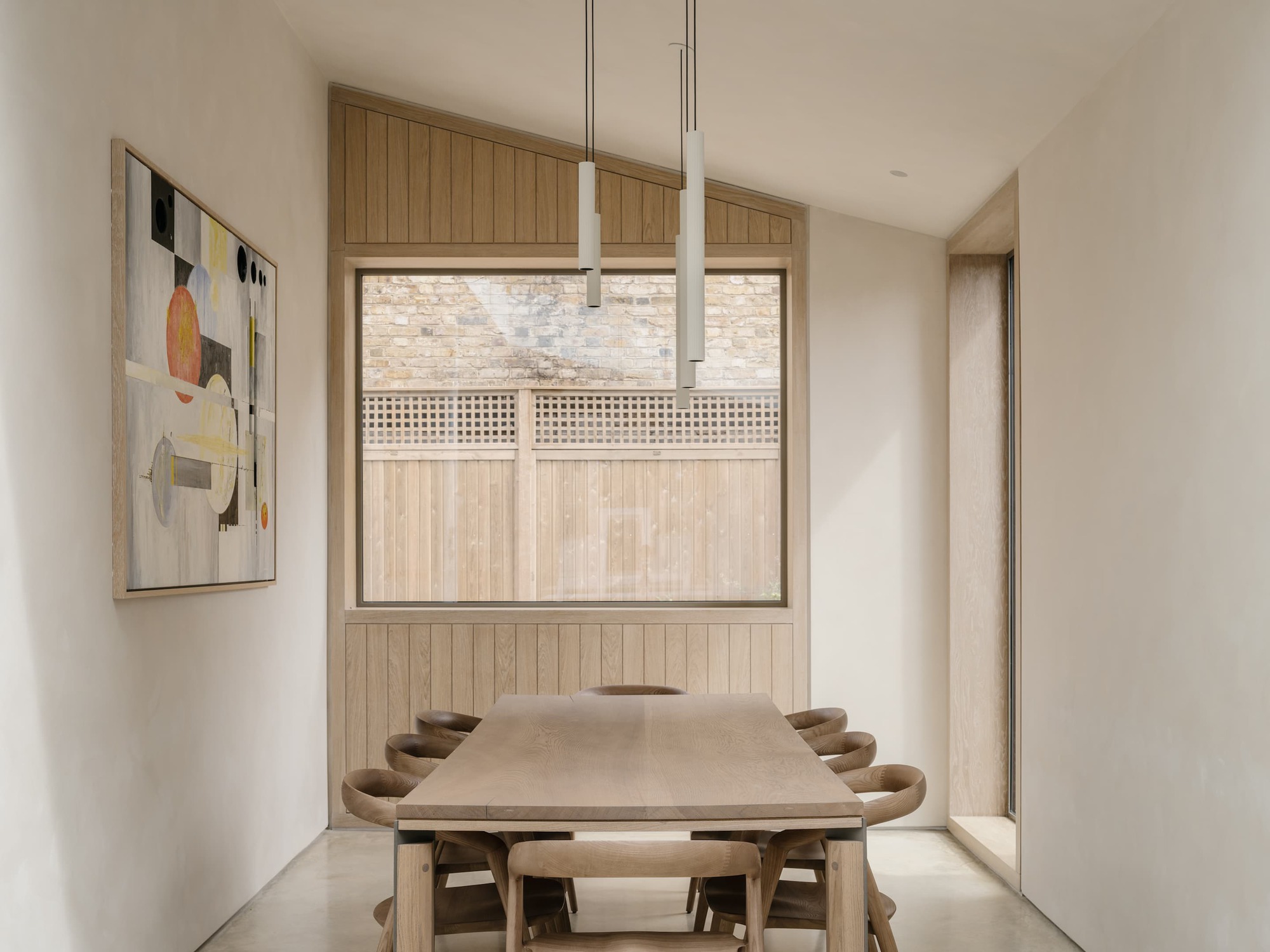 Dining room with a long oak table, pendant lights and a square window framing a timber fence and brick wall