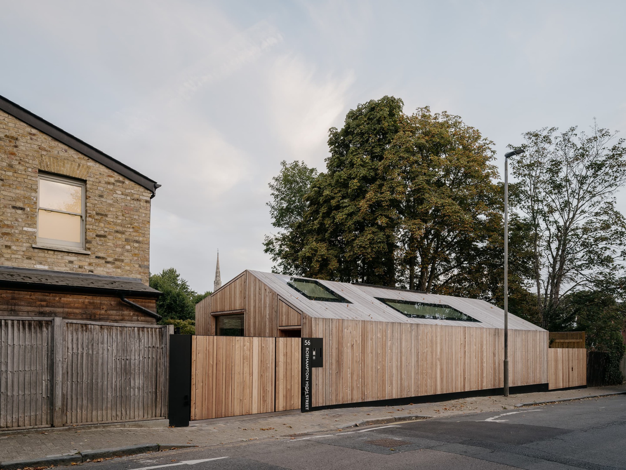 Street frontage of the timber-clad pitched volume with two rooflights behind a cedar fence, under an overcast sky with tall trees behind
