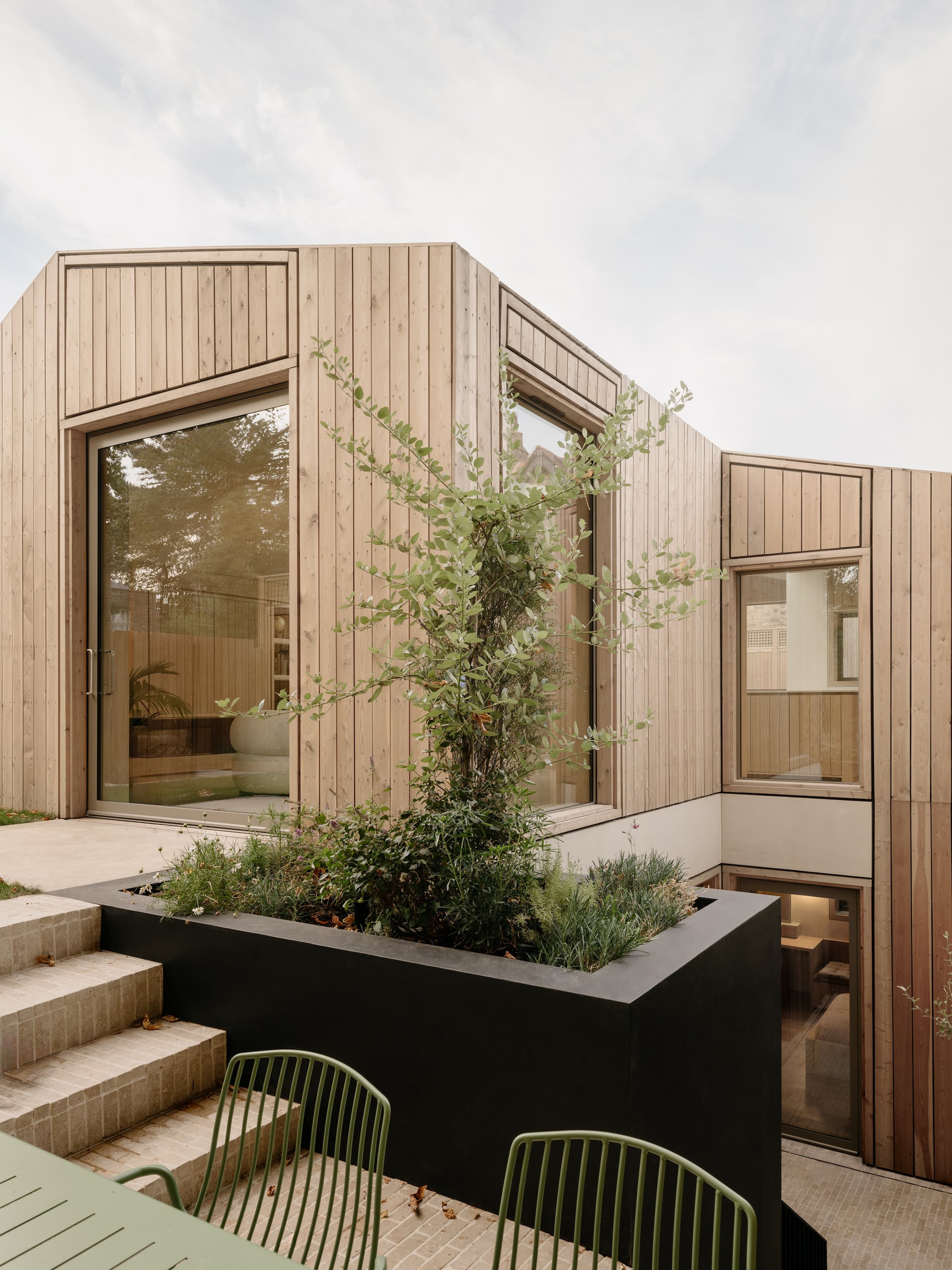 Black steel planter with a climbing shrub in front of the vertical timber-clad walls and tall glazing, beside brick steps and green chairs