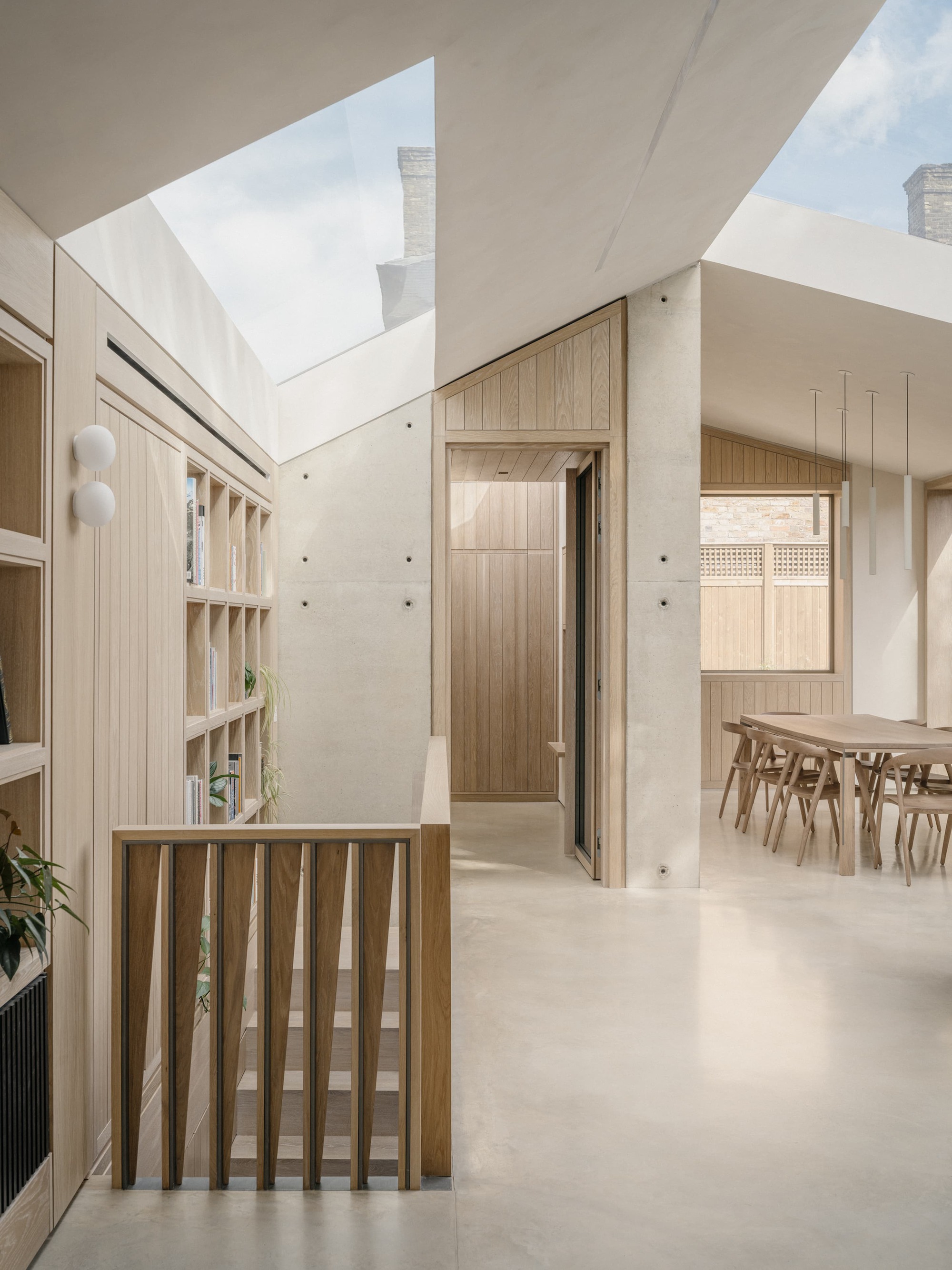 Double-height circulation space with oak shelving, slatted stair balustrade and board-marked concrete wall, lit by angled rooflights and a view of chimneys