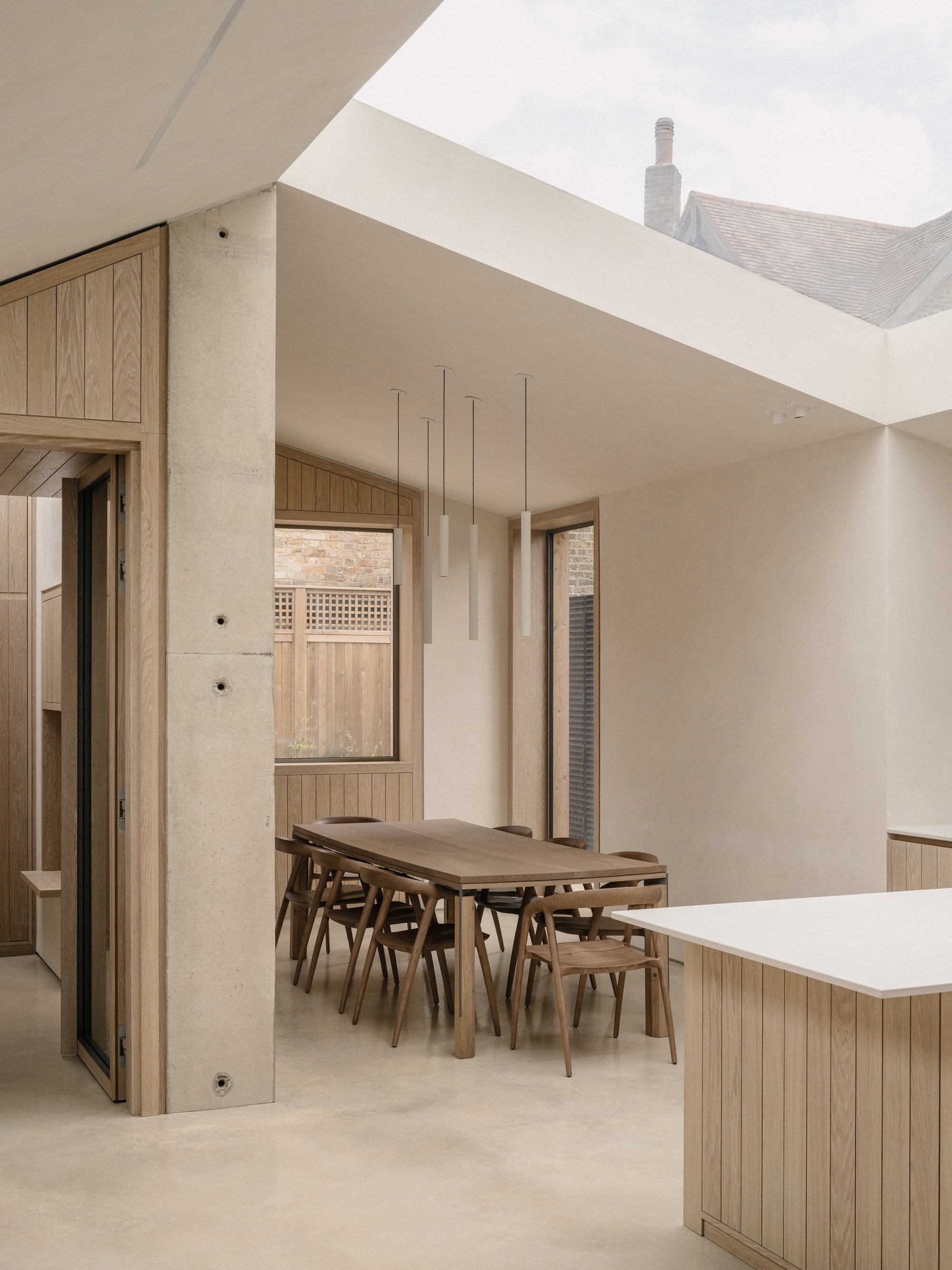 Dining table and oak chairs beneath a rooflight framing a neighbouring chimney, beside a concrete column and timber-framed doorway