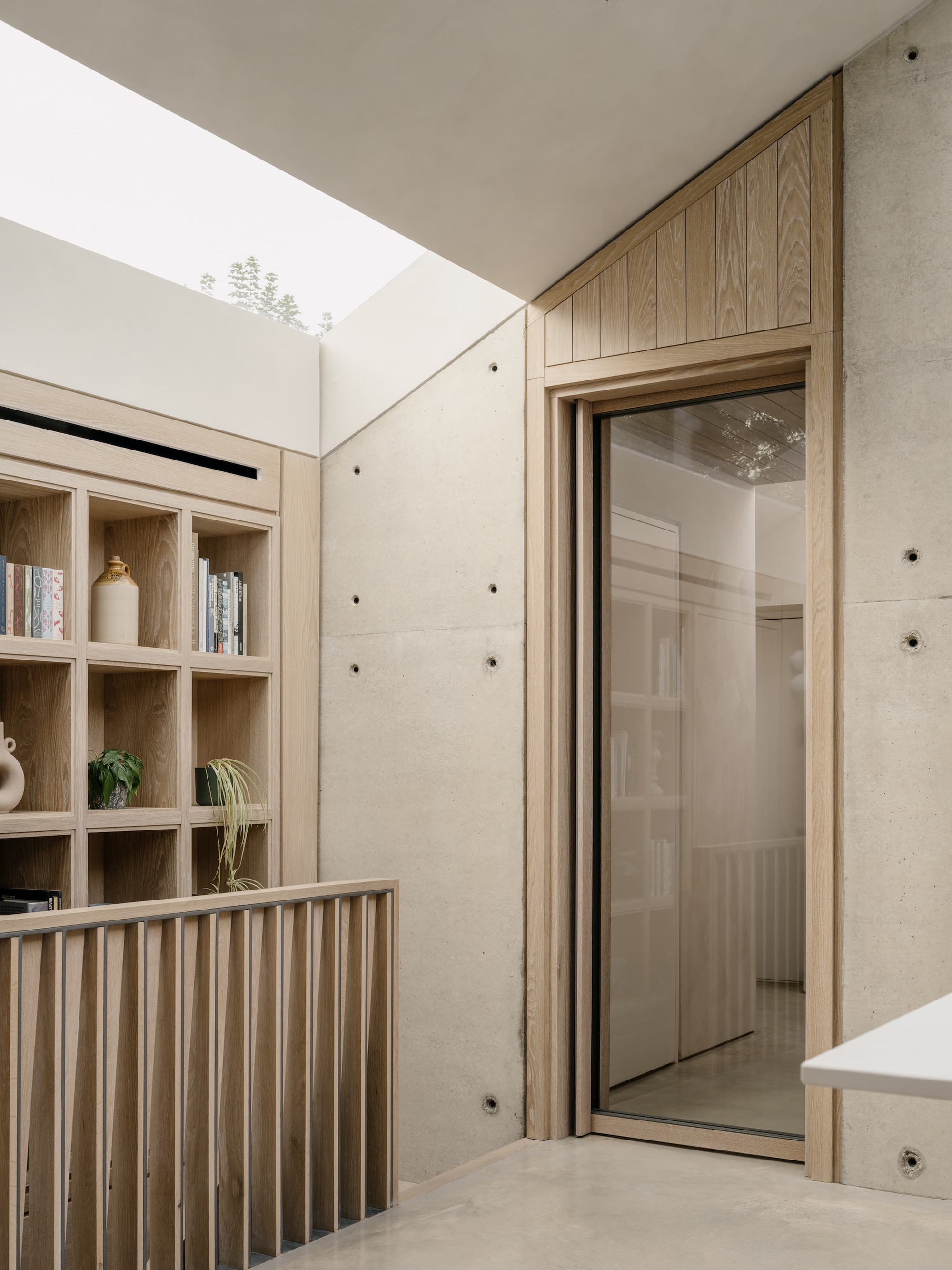 Landing with oak shelving, slatted balustrade and tie-hole concrete wall beside a glazed timber door, with daylight from the rooflight above