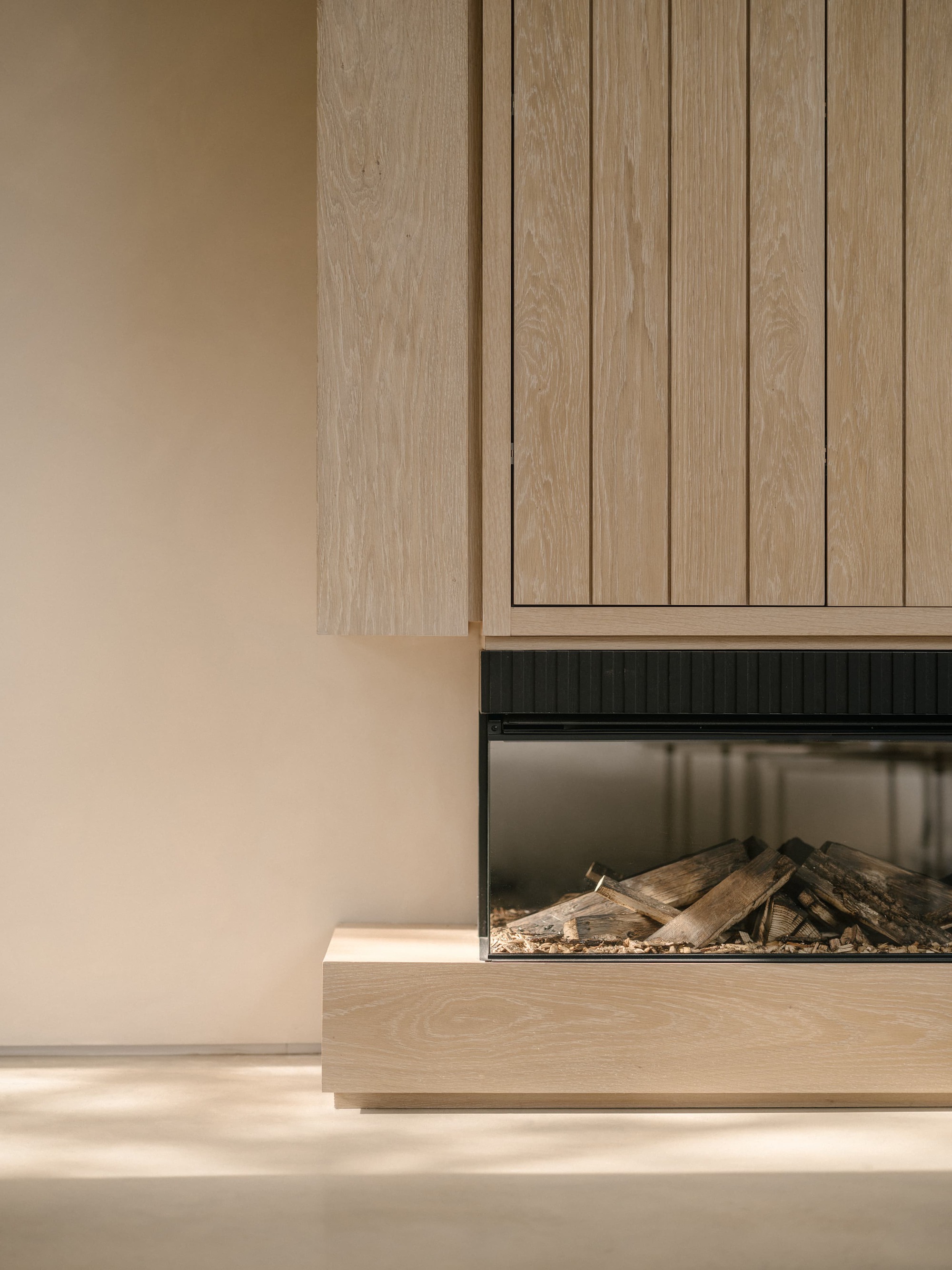 Close-up of a linear fireplace with stacked logs set into an oak surround, with soft sunlight across the floor