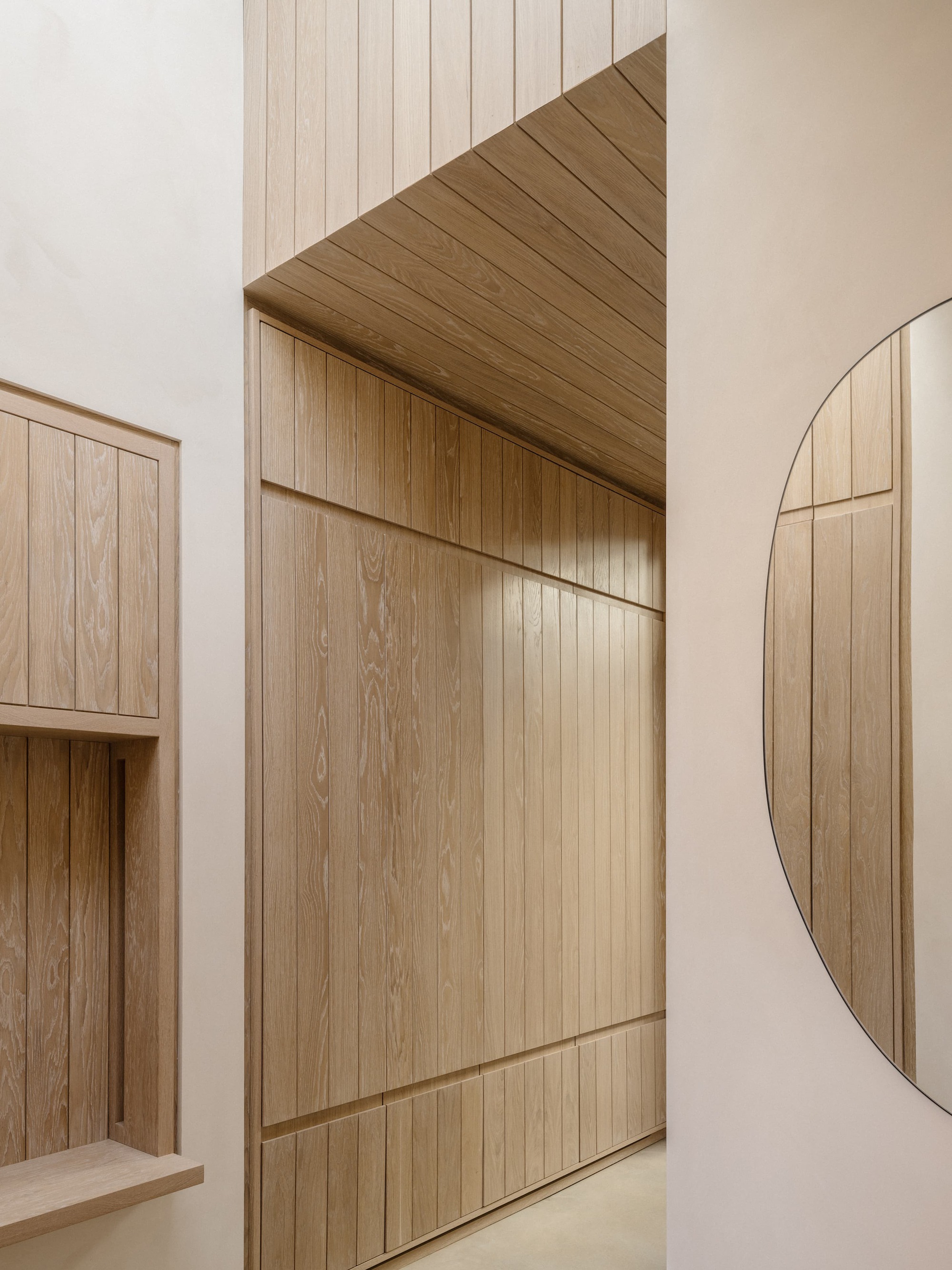 Corridor lined in vertical oak boards with a recessed shelf niche and a round mirror reflecting the paneling