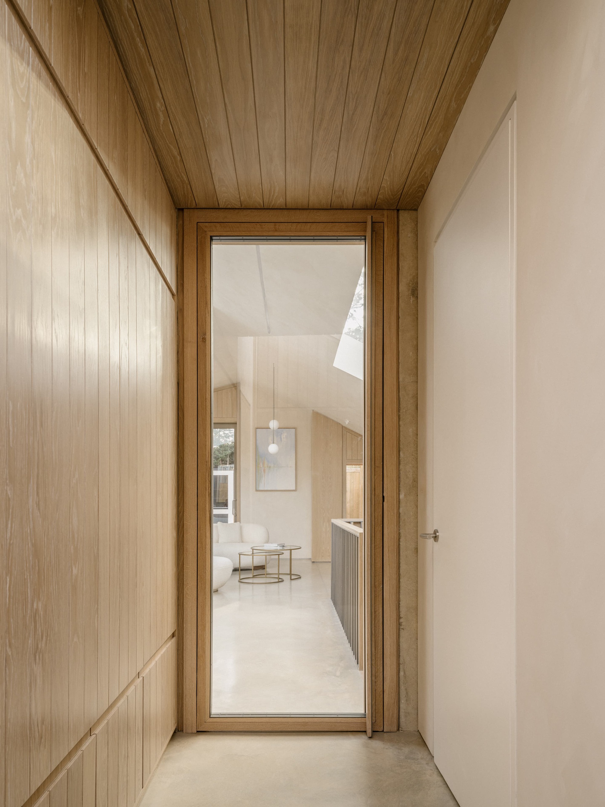 Timber-lined passage ending in a glazed oak door that frames the living room and its pendant lights beyond