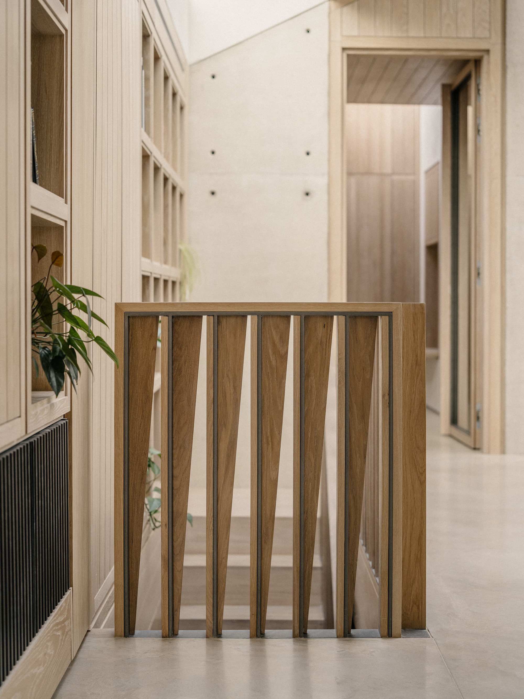 Oak stair balustrade of tapered vertical timber blades beside fitted shelving and a board-marked concrete wall in soft daylight