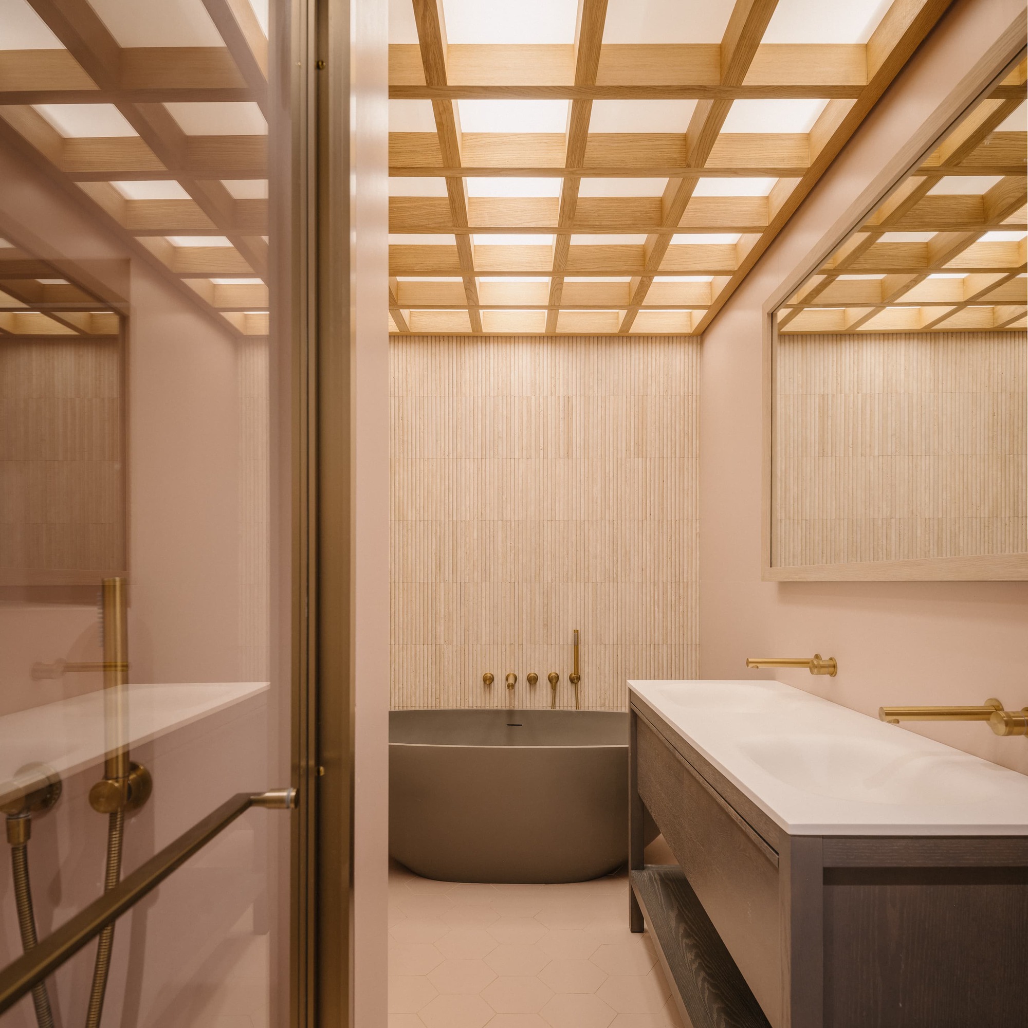 Bathroom with a timber lattice ceiling glowing with skylight above a grey freestanding tub, brass taps and a pink wall