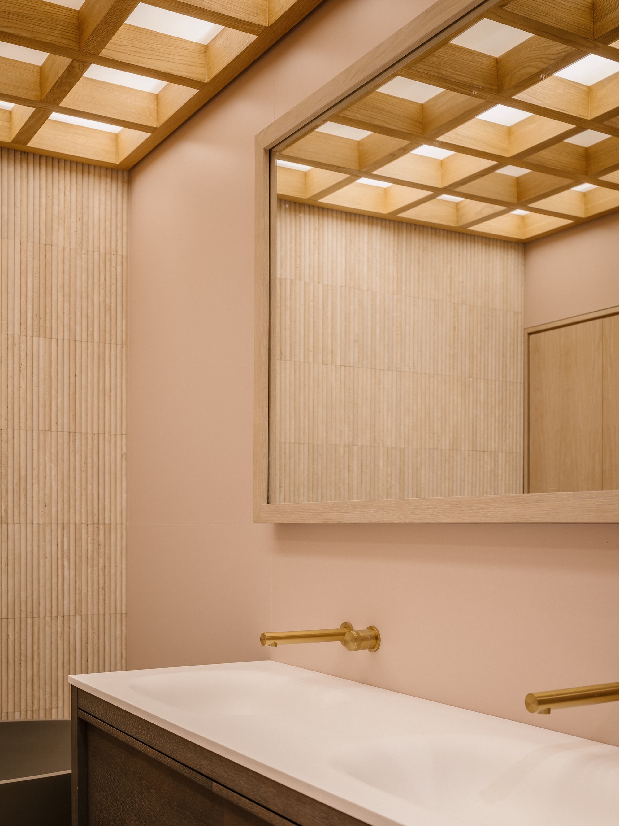 Double basin vanity with brass wall taps, fluted stone tile and a mirror reflecting the glowing timber grid ceiling