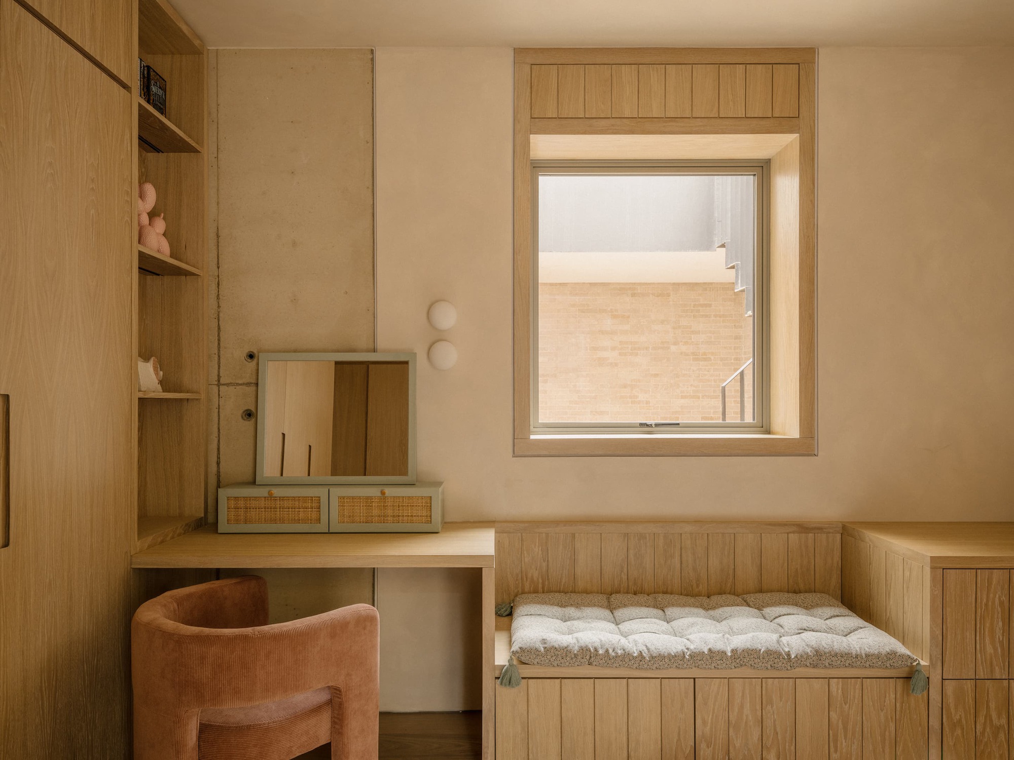 Bedroom corner with a built-in desk, rattan drawers, window seat with tasselled cushion and a square window onto a brick wall