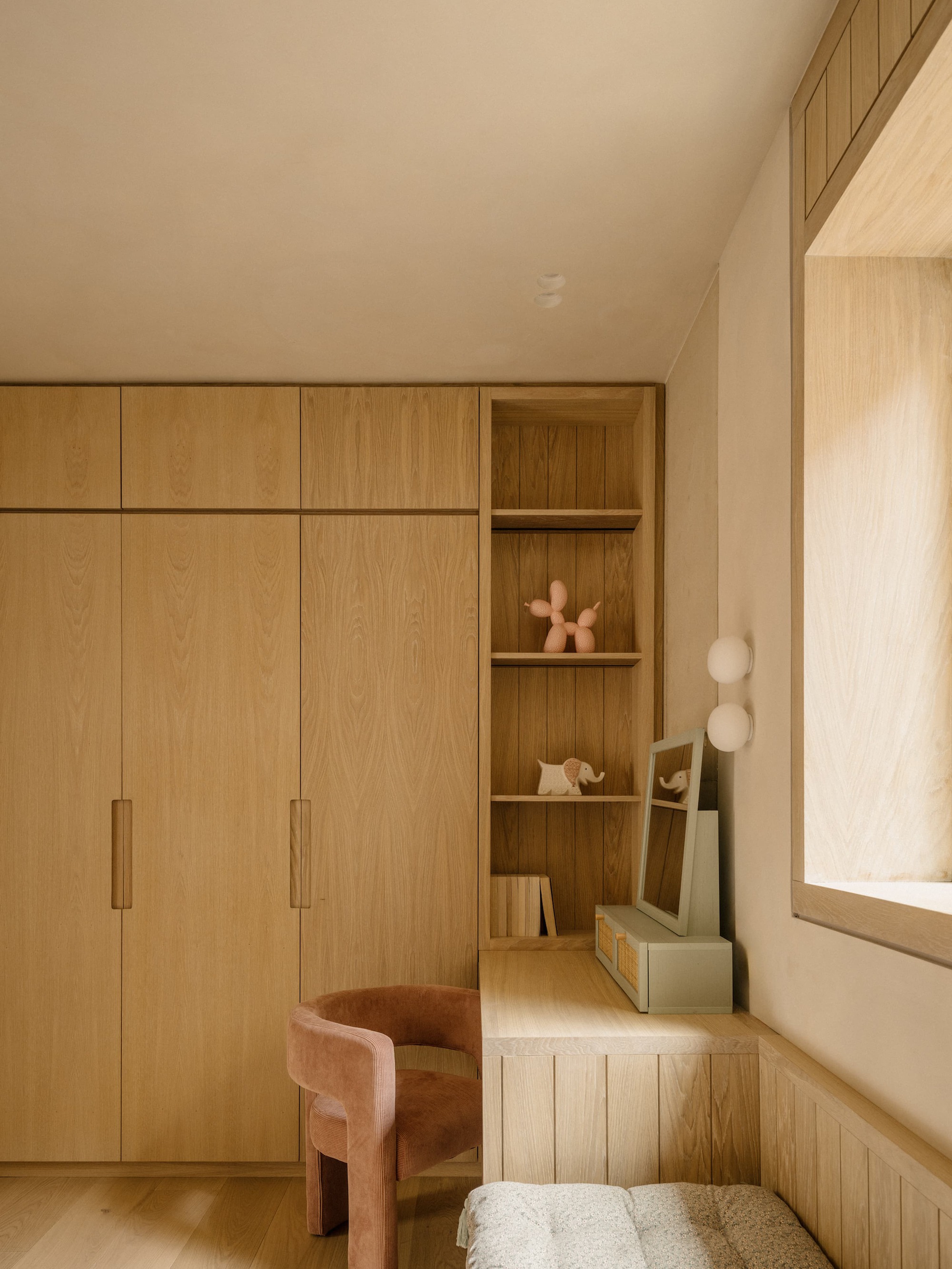 Oak wardrobe doors beside open shelving with balloon-dog figure, a rust velvet chair and a deep window reveal in daylight