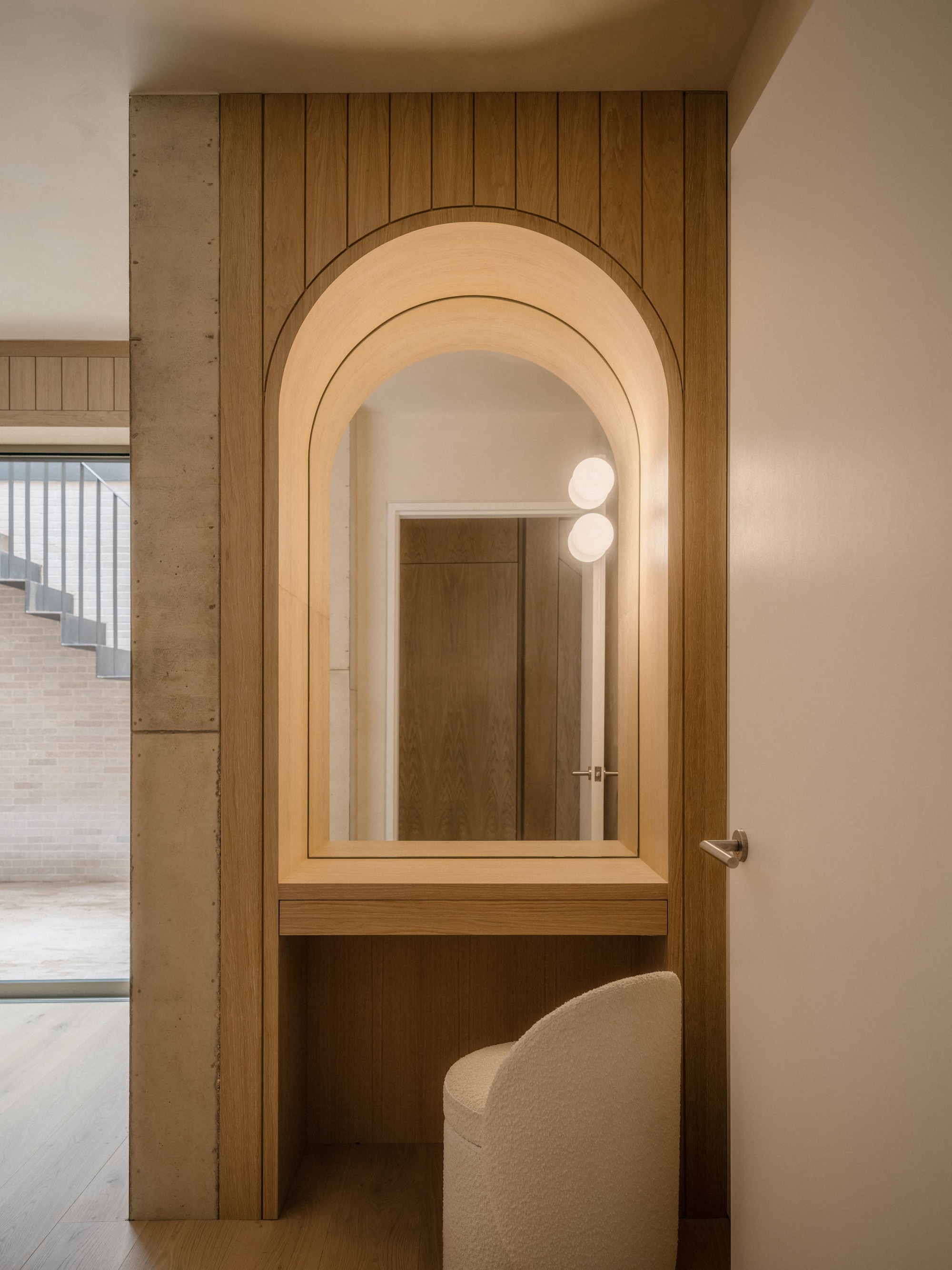 Arched oak-framed mirror and built-in dressing shelf beside a board-formed concrete column, with a boucle stool in warm light