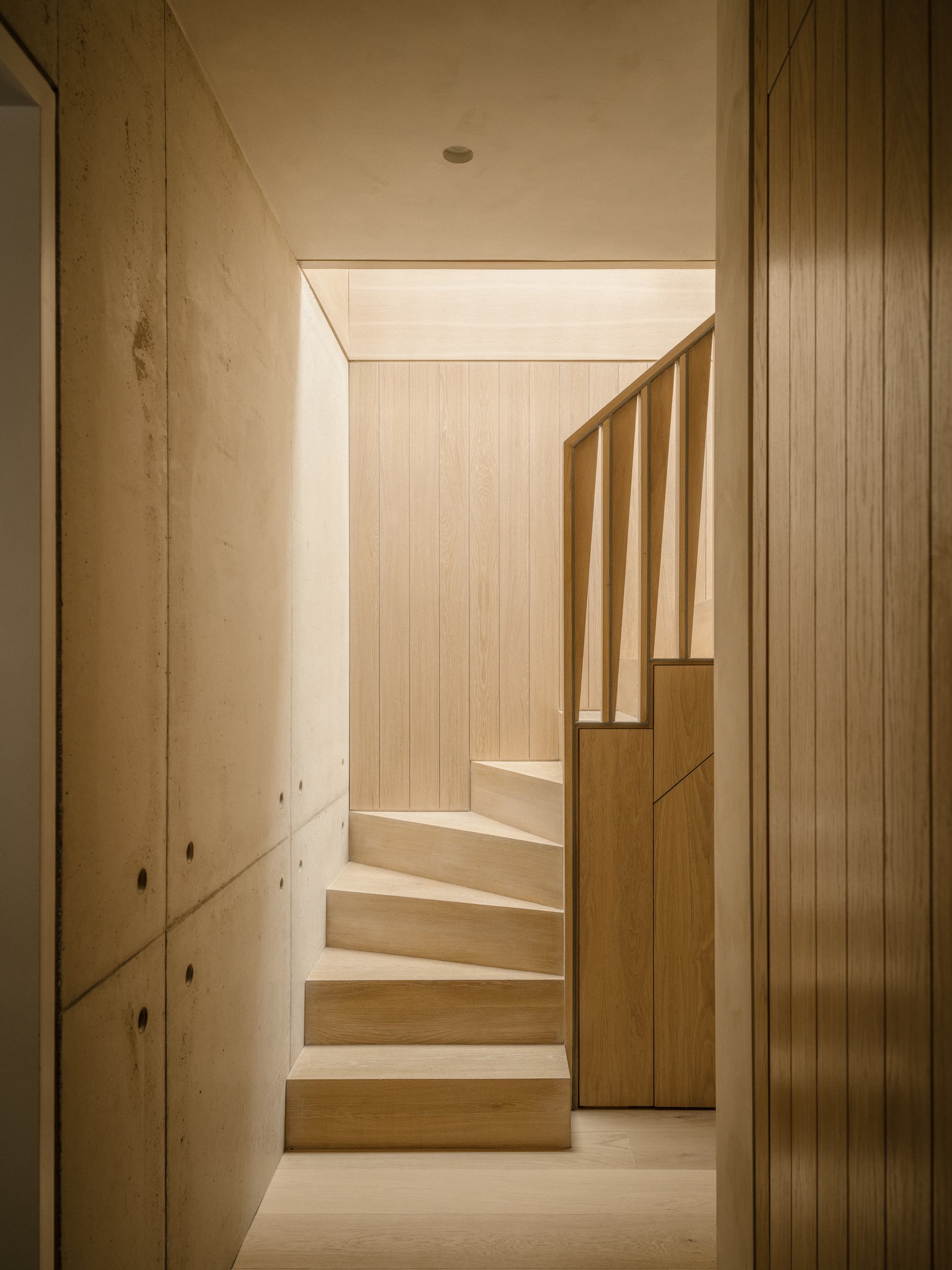 Winding oak staircase with angled balustrade between board-marked concrete walls, lit by a soft skylight glow from above