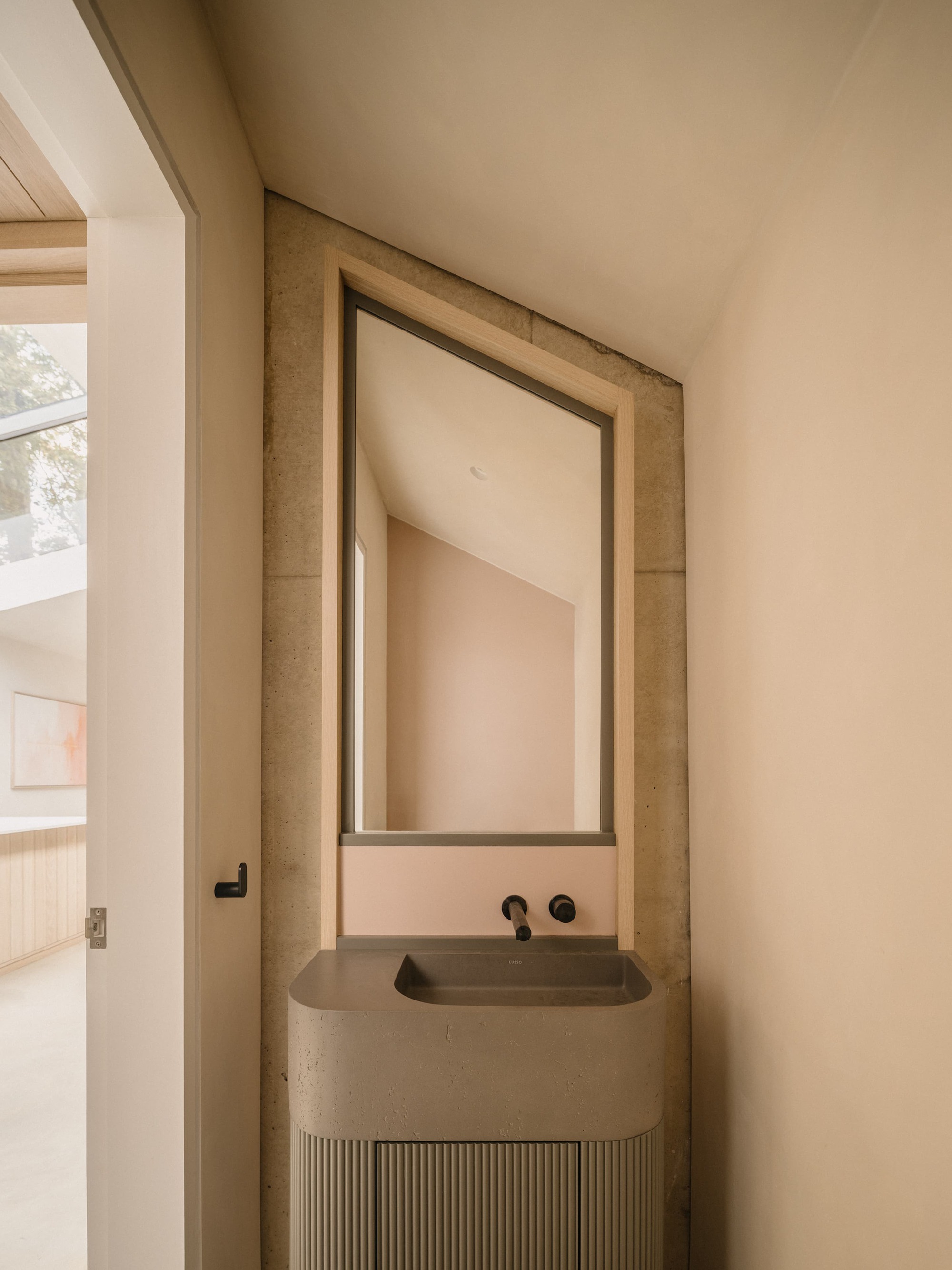 Concrete washbasin with fluted cabinet beneath a timber-framed mirror on a sloped ceiling wall, with pink tiled splashback