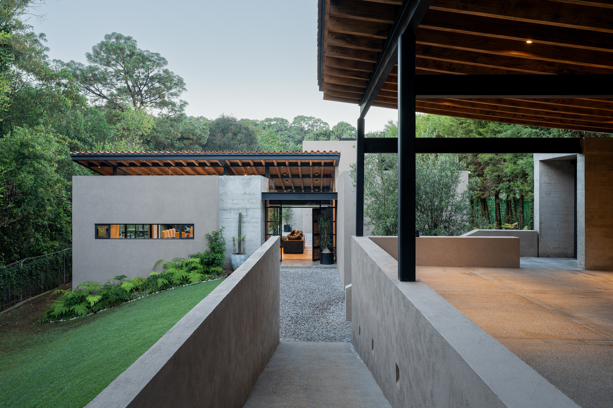 Gravel path between two concrete walls leading to a glazed entry, with exposed timber rafters on steel columns at right