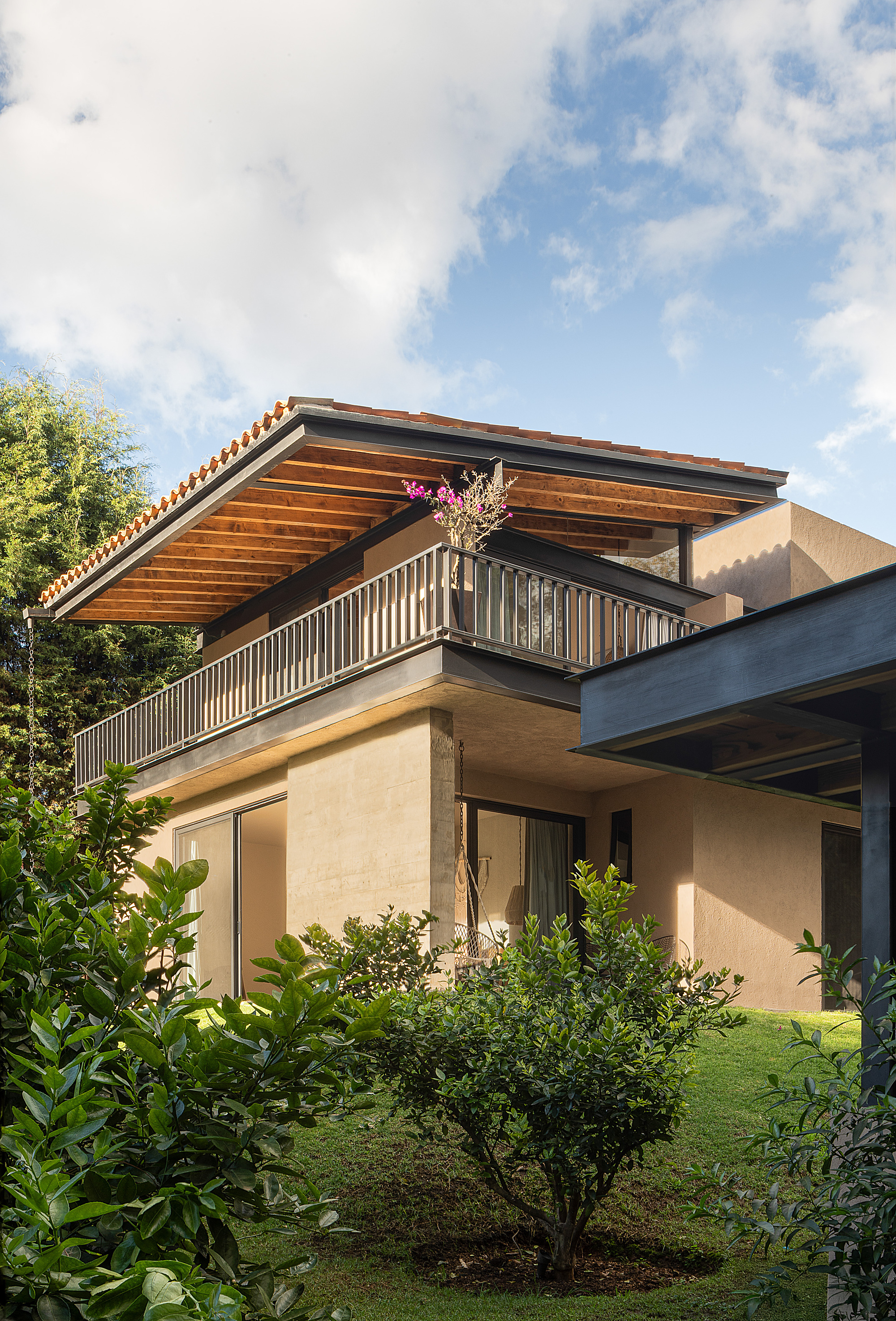 Upper terrace with steel railing under a clay tile roof and timber soffit, rising above rendered walls and shrubs in afternoon sun