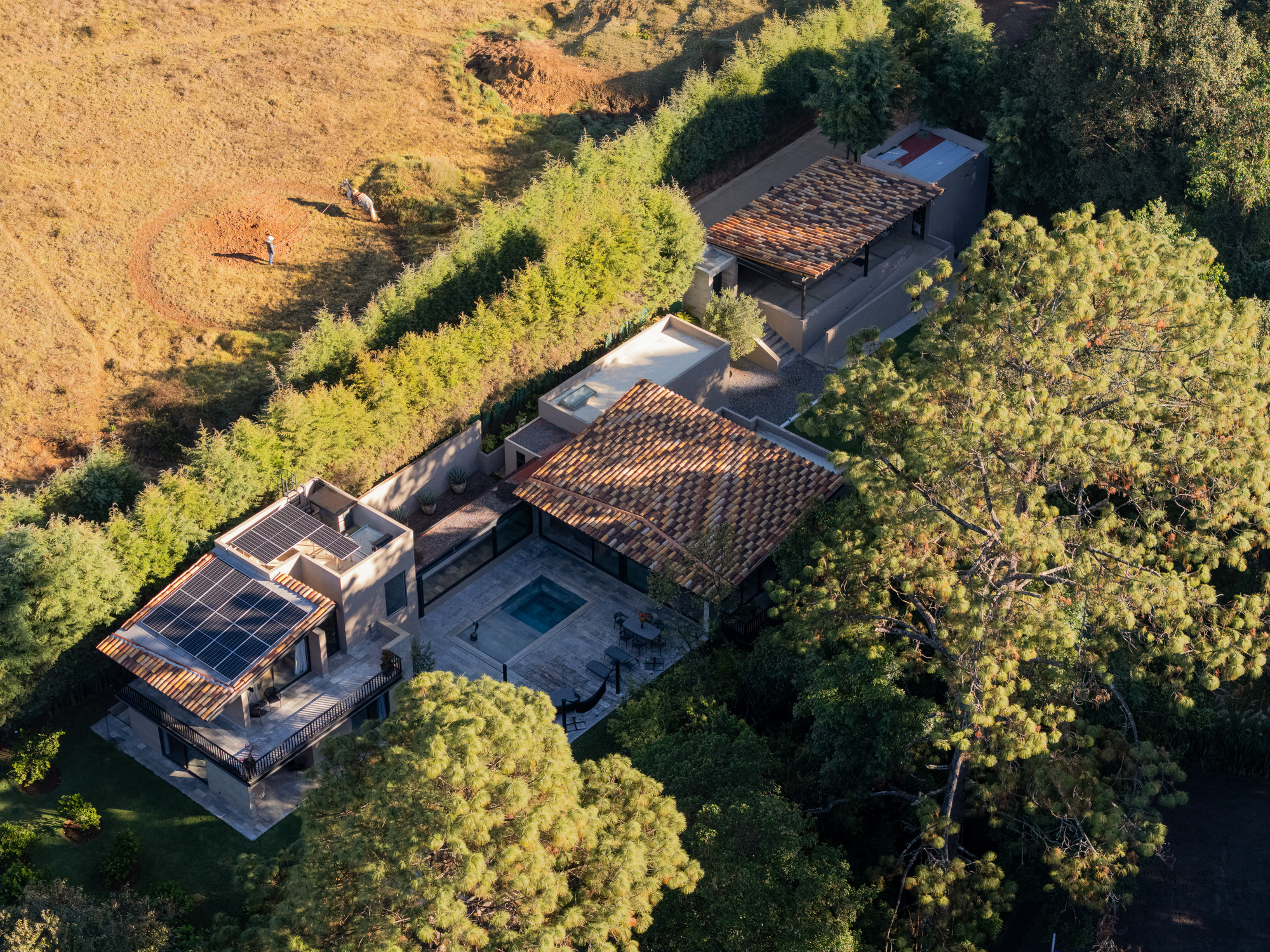 Aerial view of clay tile roofs, a solar panel array and a courtyard pool among pine trees in low golden light