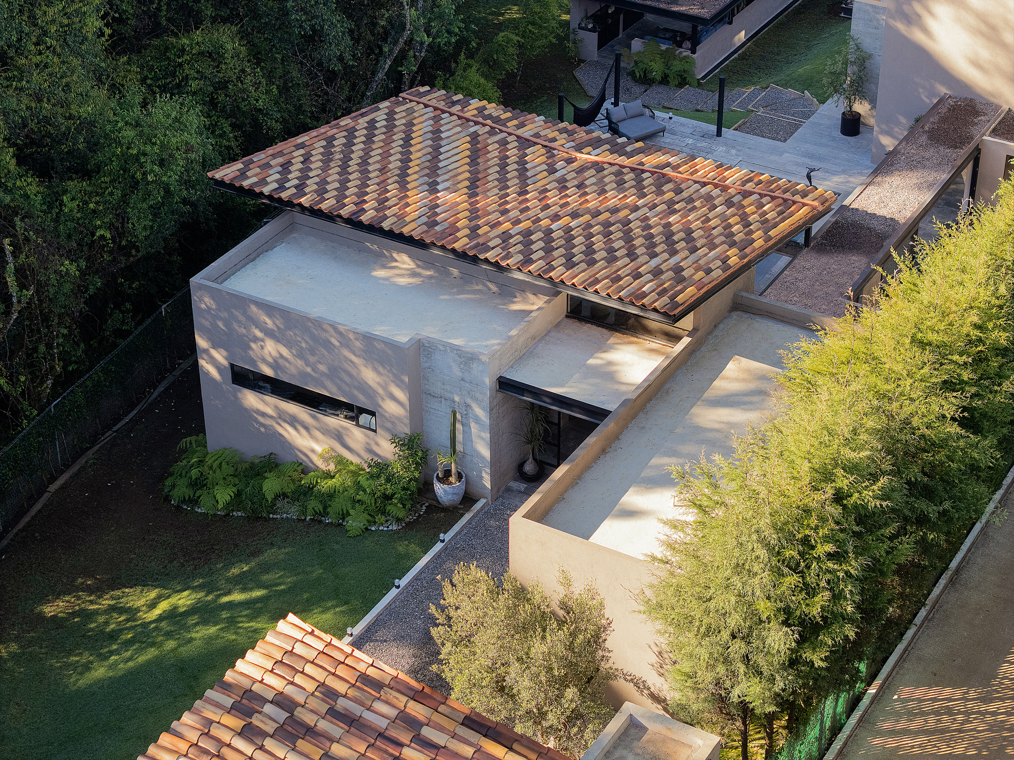 Elevated view of a pitched multi-toned clay tile roof over rendered volumes with a flat terrace, shaded by trees