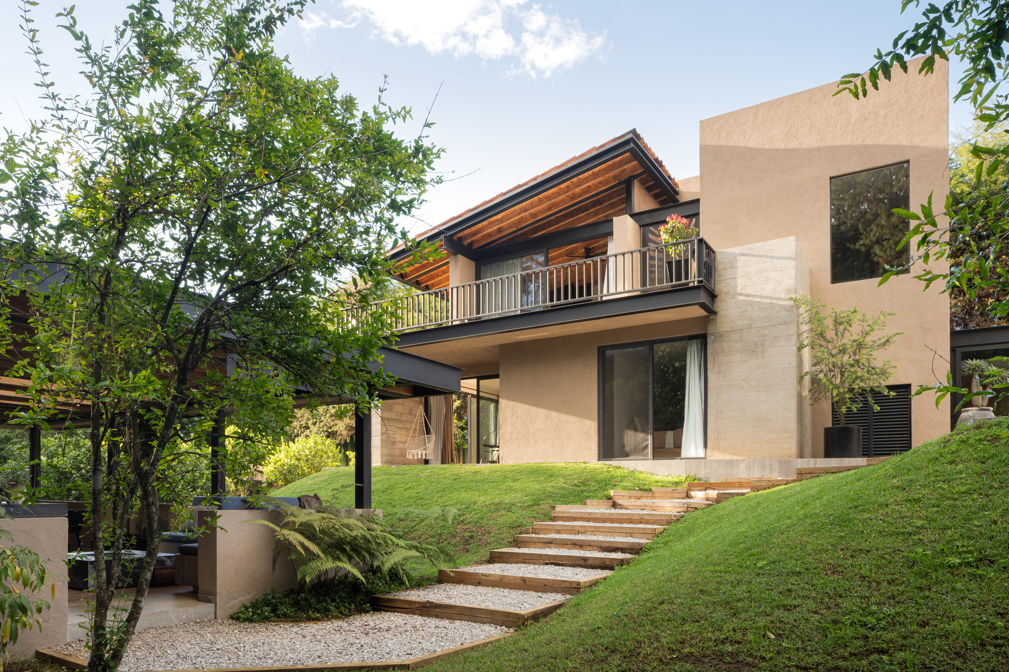 Rendered house facade with a balcony and timber eaves above a grass slope and gravel steps with timber risers