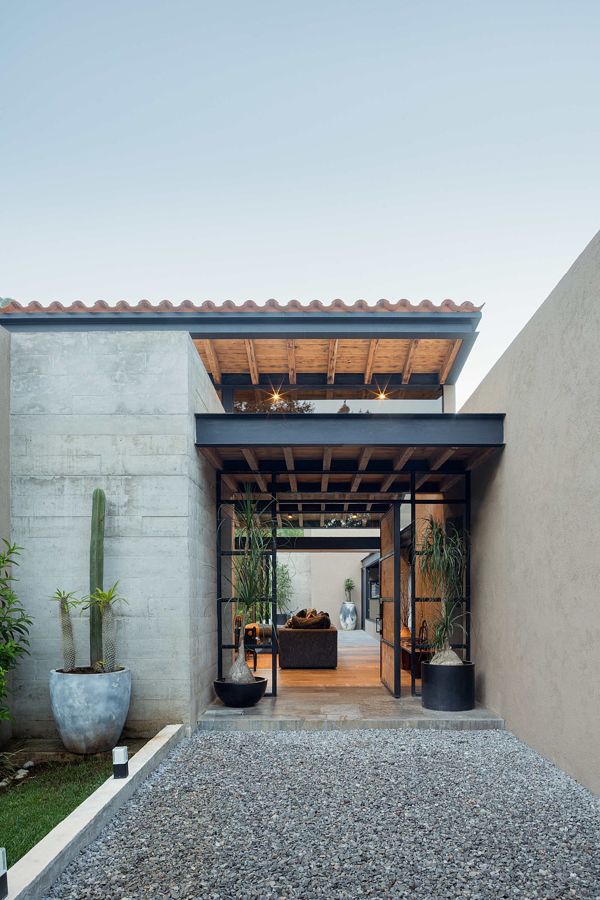 Entry passage framed by steel pivot doors and a board-formed concrete wall, with potted cactus and gravel at dusk