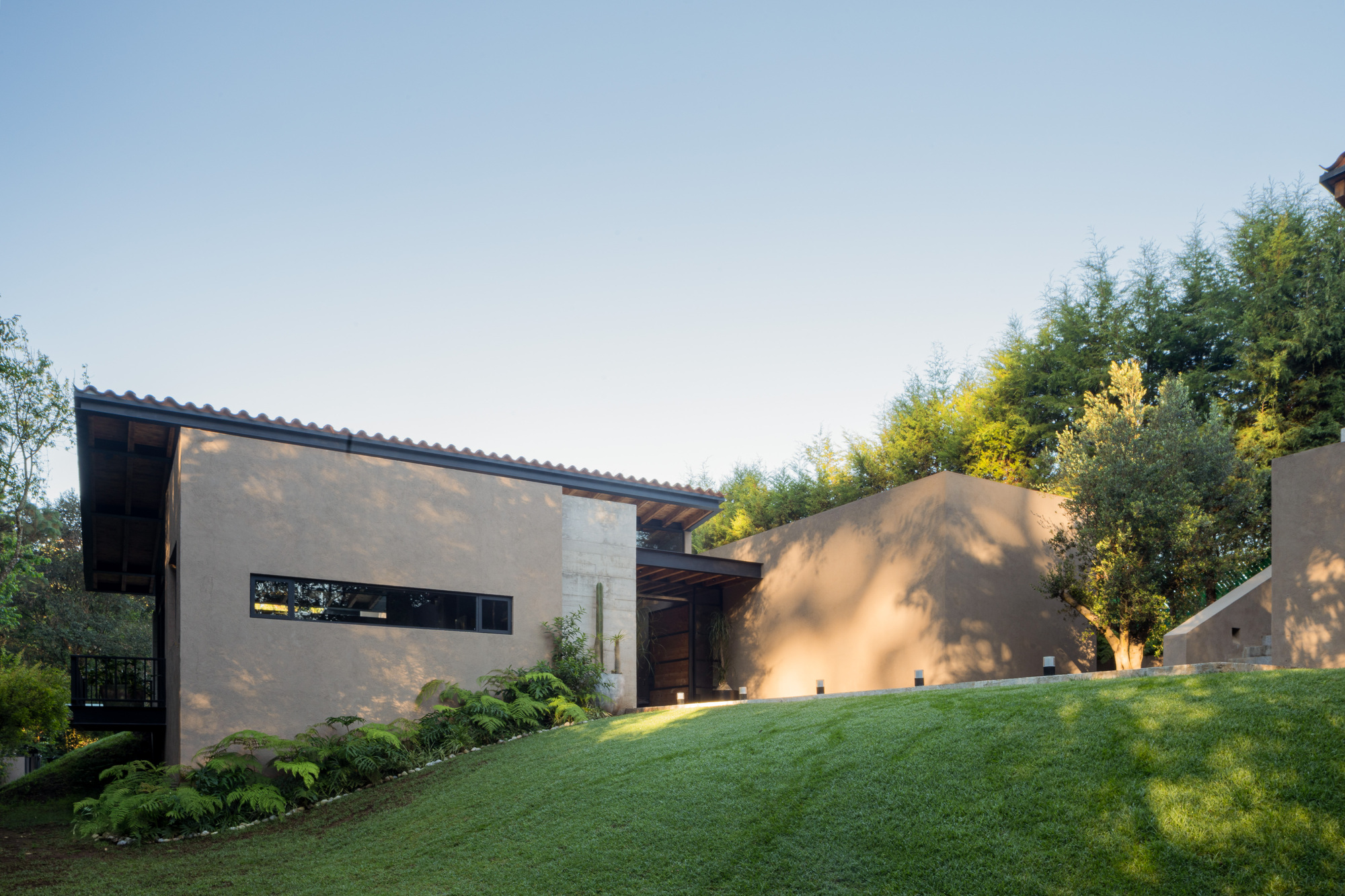 Stucco facade with a long ribbon window and tiled roof, set behind a grassy slope in low afternoon sun