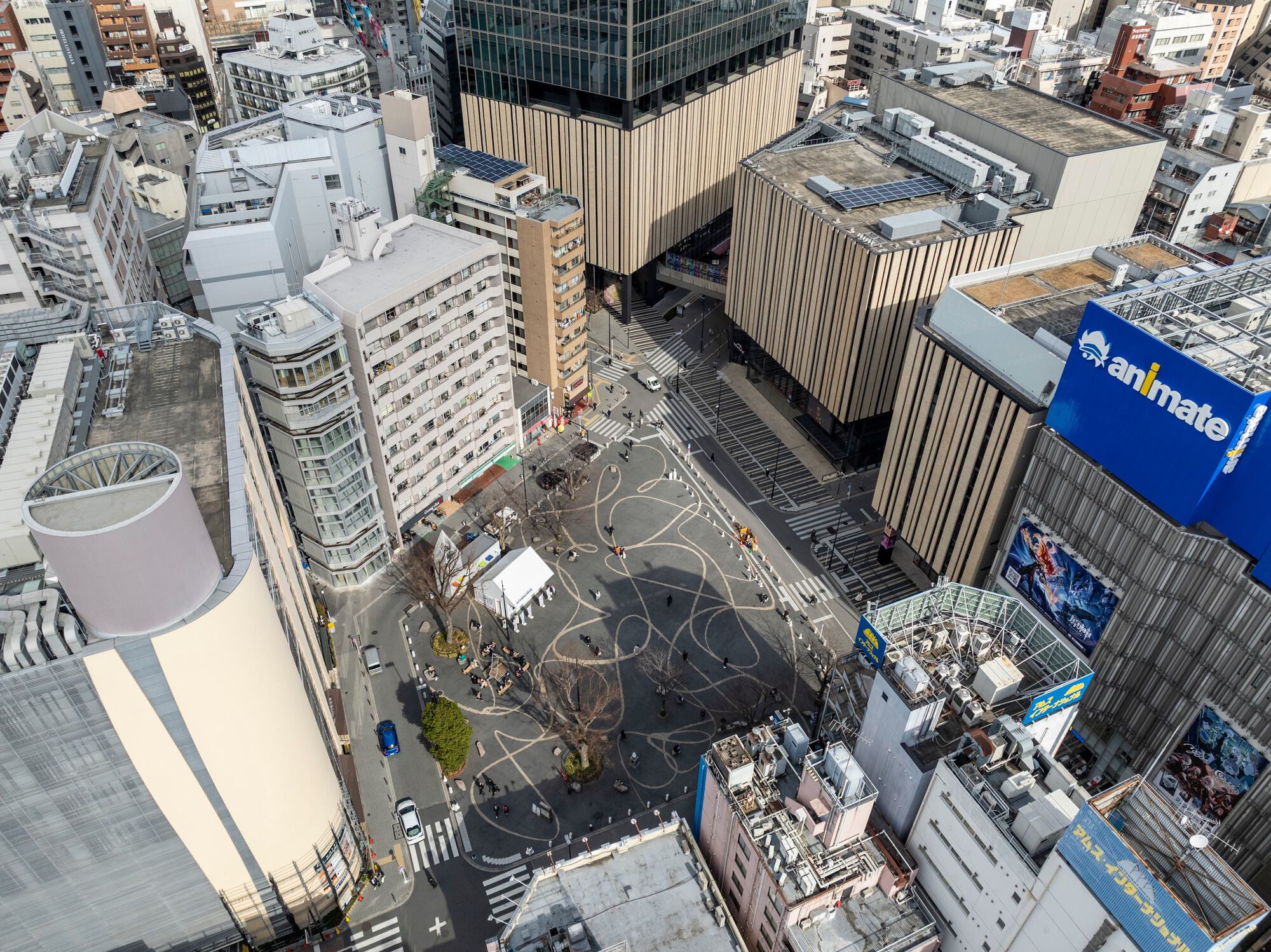 Aerial view of a plaza with curving painted line patterns, surrounded by buildings with vertical timber-toned fins and a large animate sign
