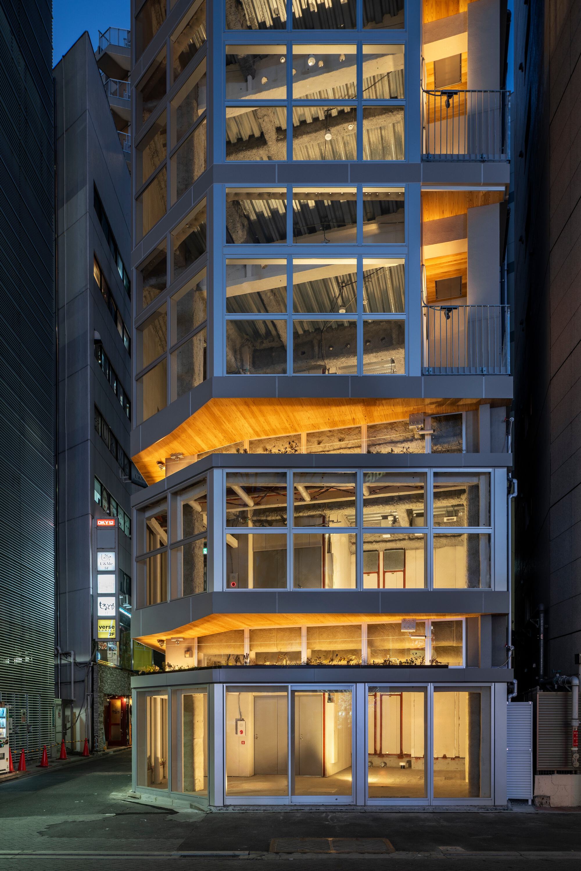 Narrow tower facade at night, with glowing timber soffits and glazed floors showing exposed structure above a ground-level shopfront