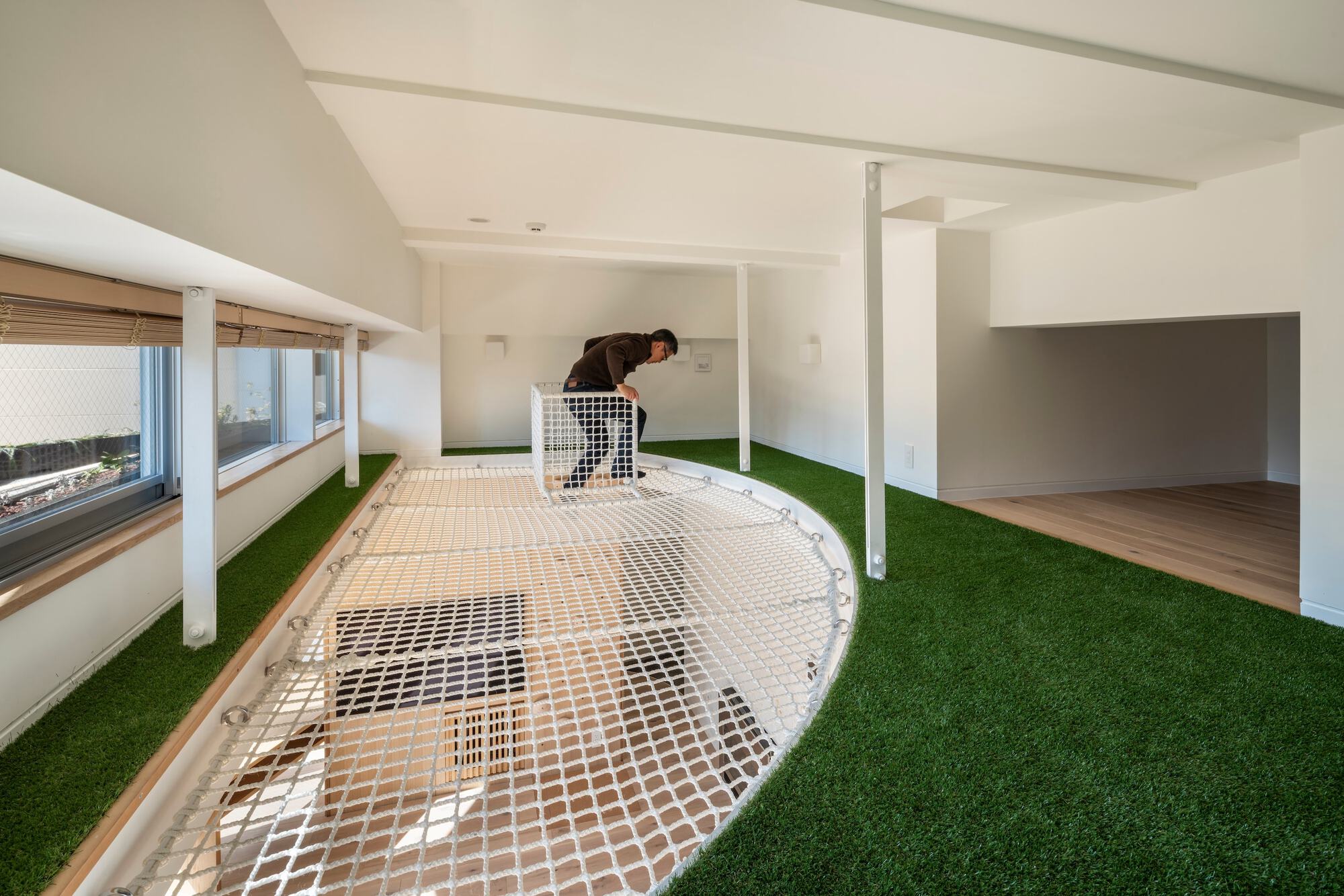 Loft room with white rope net floor set into artificial turf, a person crouching beside a net-fenced opening
